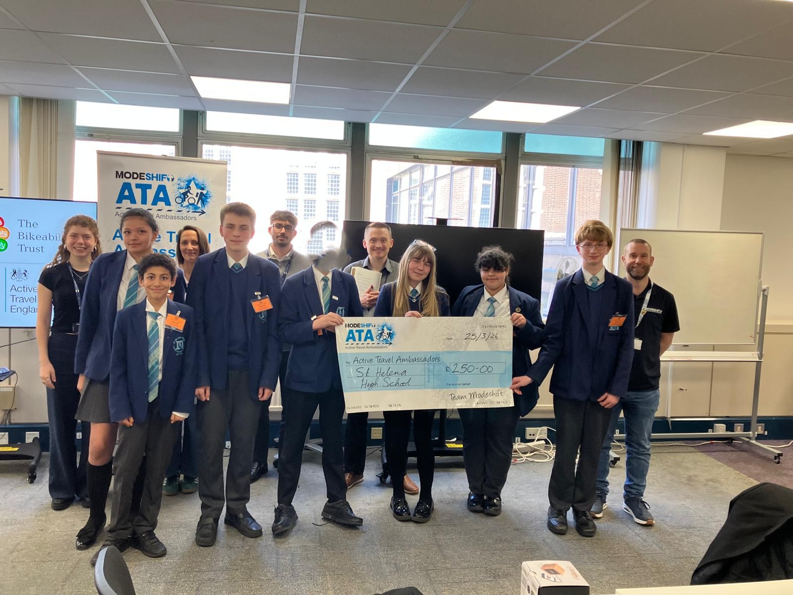 Group of students and adults in a classroom holding a large check for £250. Active Travel Ambassadors branding on the check.