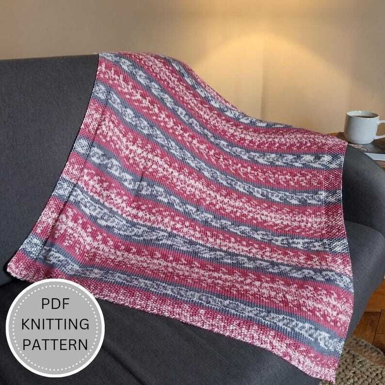 A pink, grey and white striped knitted blanket is draped over a dark grey sofa. A circular text logo reading “PDF Knitting Pattern” appears in the lower corner. A warm lamp glows on a beige wall, with a white mug on a book beside it.