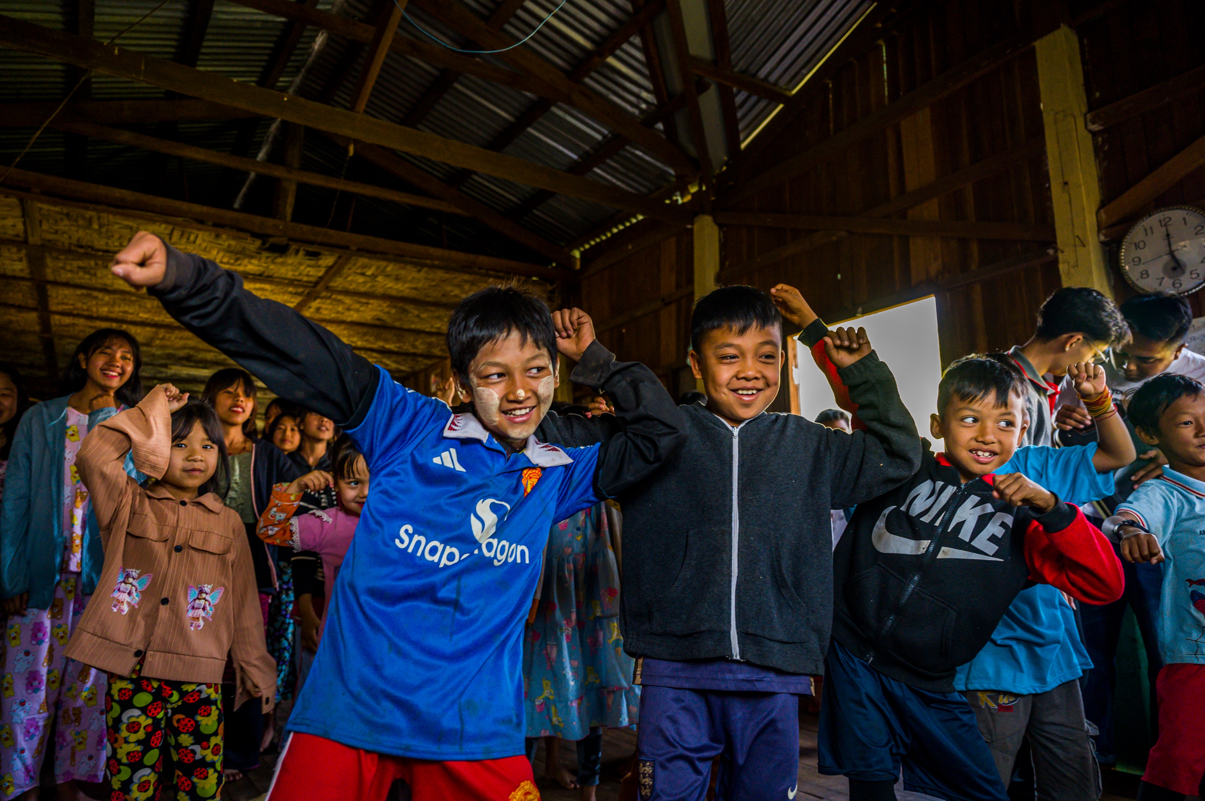 A group of young children excitedly practice a coordinated dance in a large wooden room, with one boy in the center wearing a blue jersey striking an enthusiastic pose.
