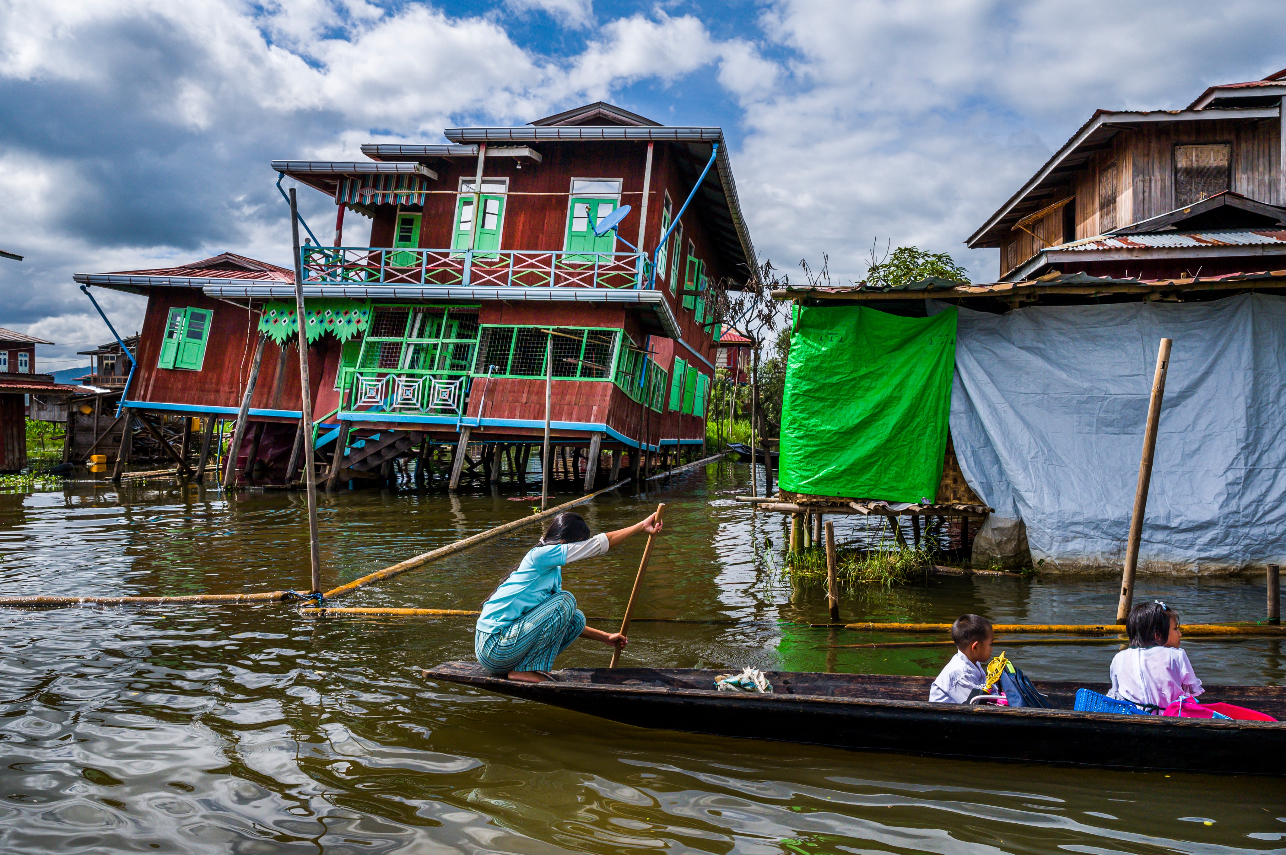 A scene on the water showing a woman rowing a traditional wooden canoe, with a small child and young girl seated in the front. Behind them are several wooden houses built on stilts over the water, including a brightly painted red and green home.
​