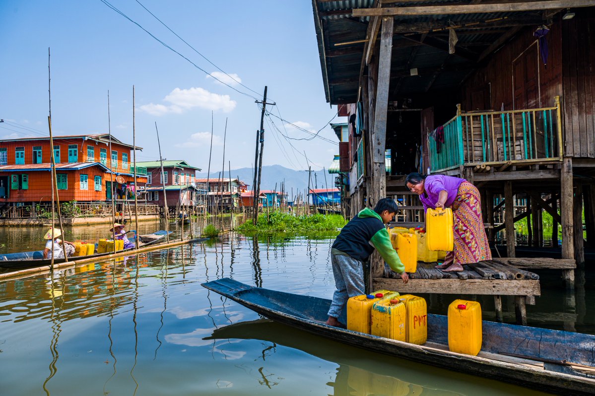 A young boy and a woman stand on a wooden platform over the water, carefully loading bright yellow water containers onto a traditional wooden boat beside their stilted houses.