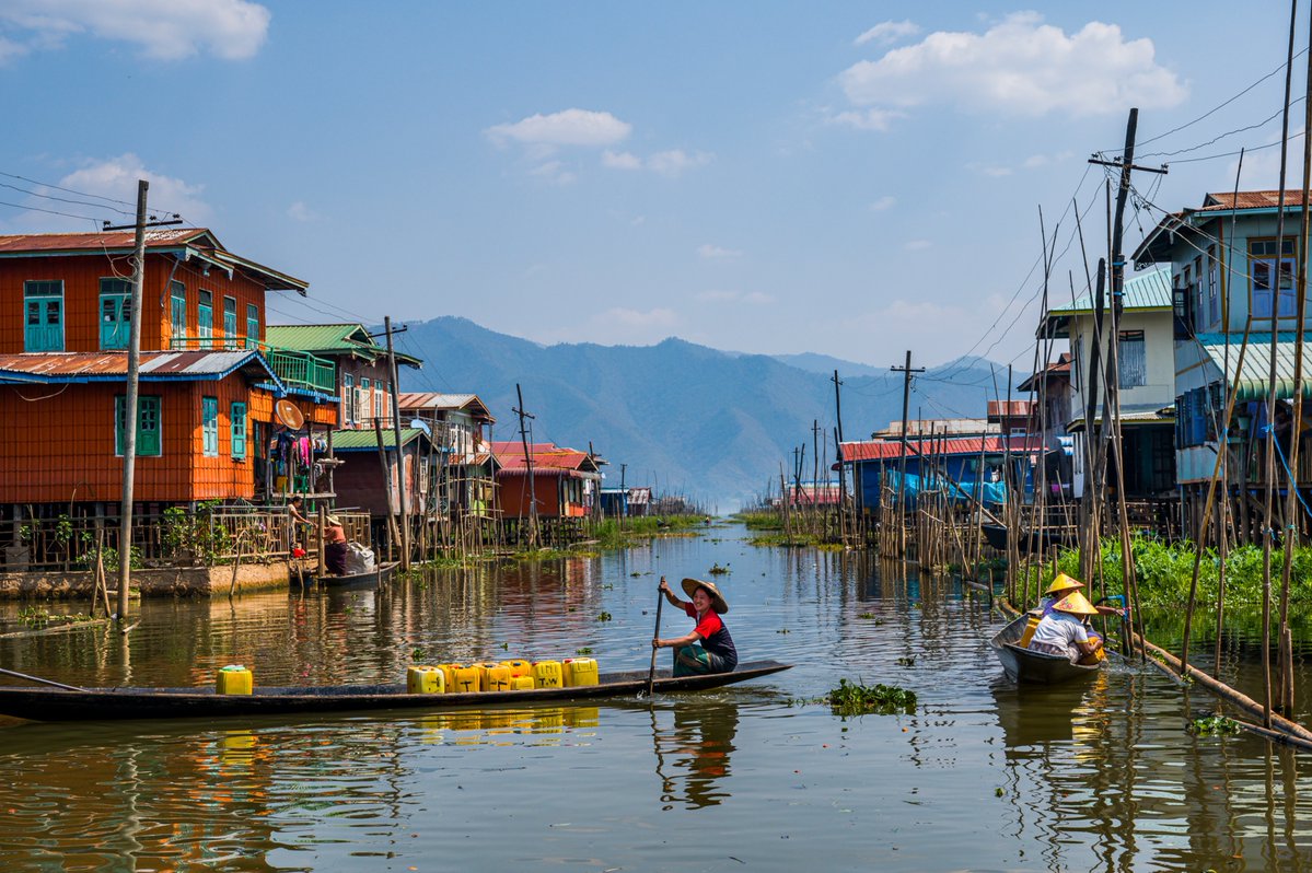 A wide view of a floating village featuring brightly colored wooden houses built on stilts, where local residents navigate the calm waters in small wooden boats against a mountain backdrop.