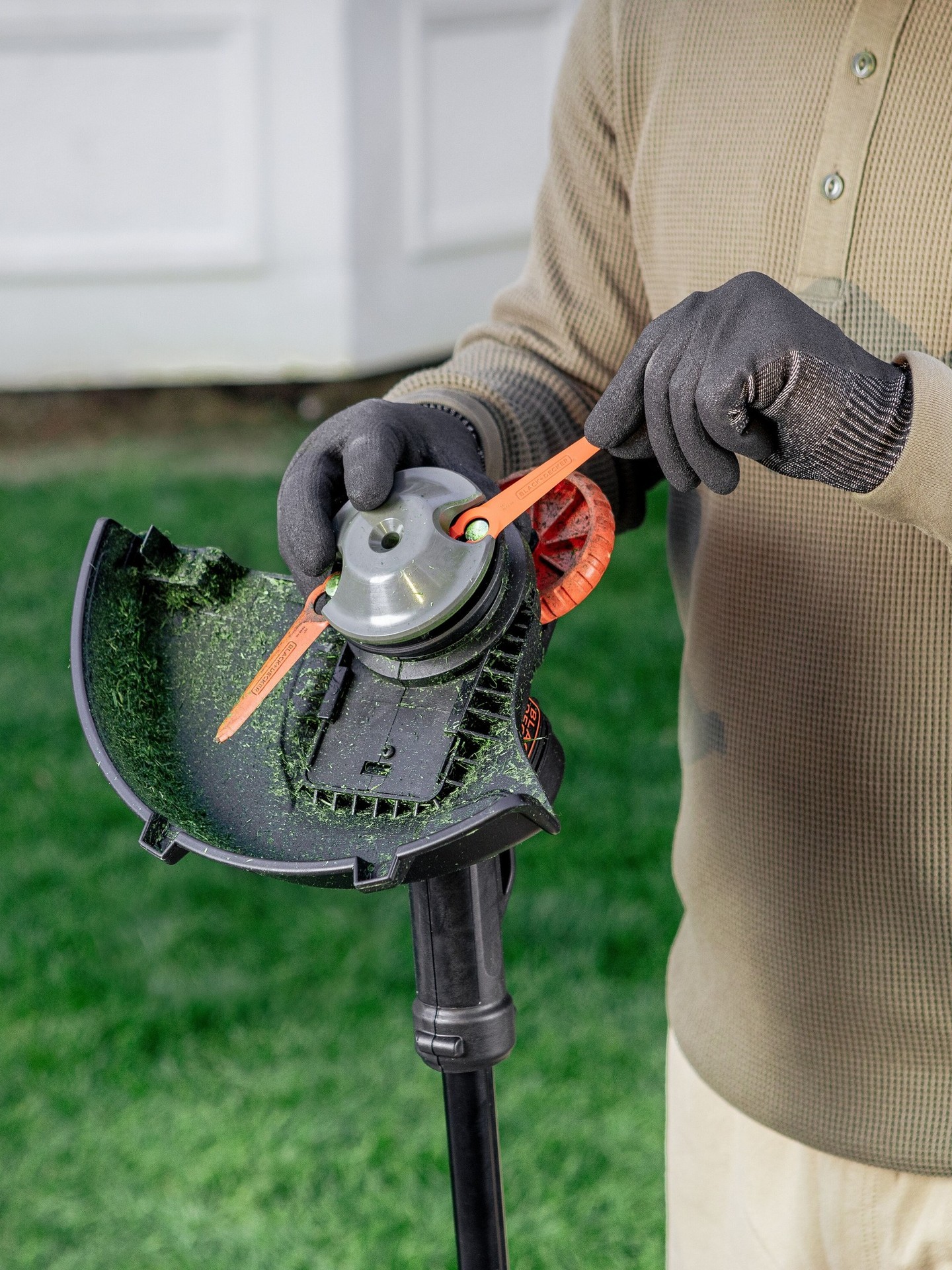 Close up of someone in gloves fixing a BLACK+DECKER@1 grass trimmer against a green lawn background. The person holds an orange tool to adjust the trimmer's head, which is partially filled with cut grass.