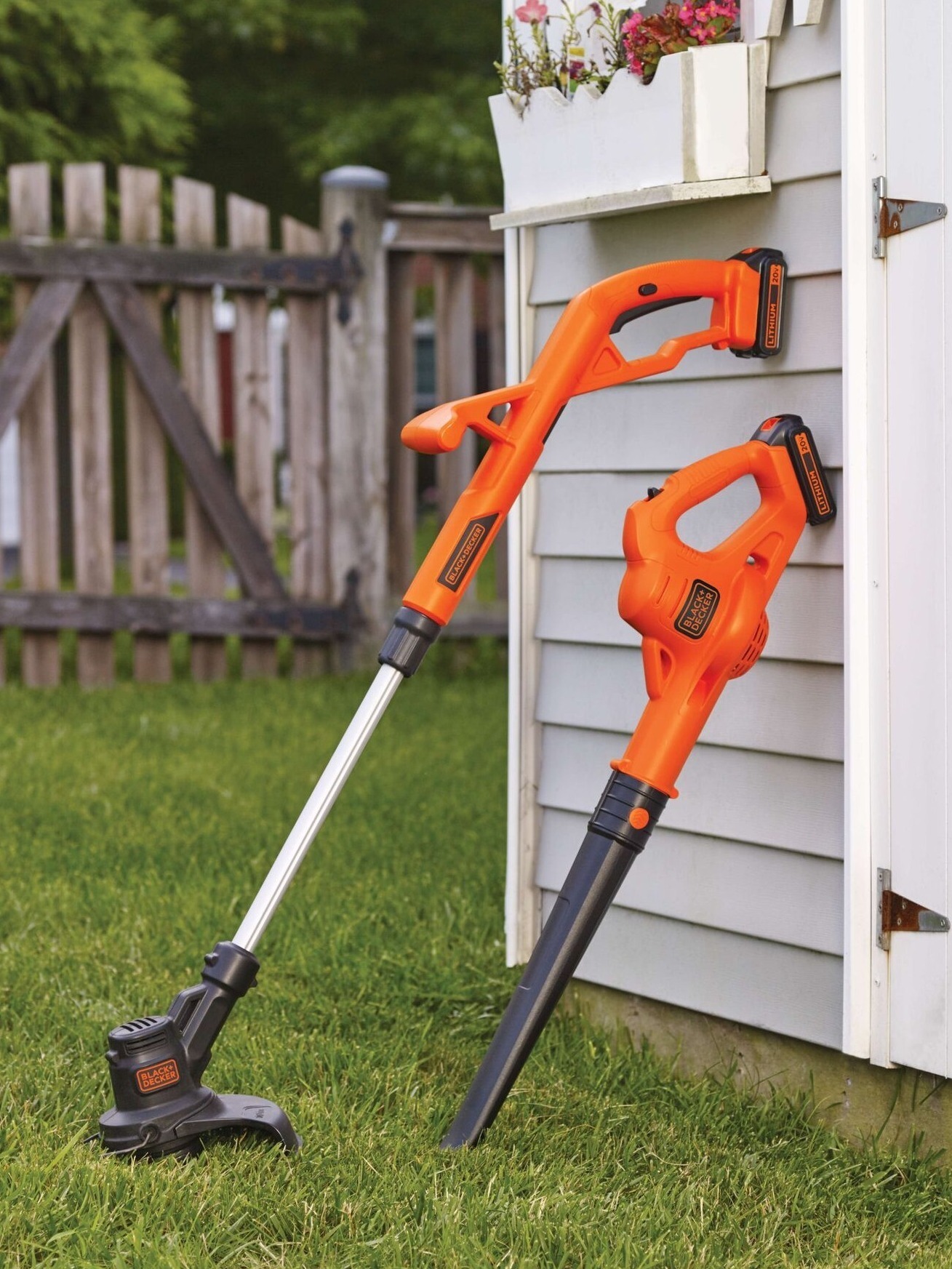 Outdoor scene featuring a Black & Decker string trimmer and leaf blower. The trimmer stands on the lawn. The blower is against a wall, both are orange & black. A wooden fence and flowers add to the background.