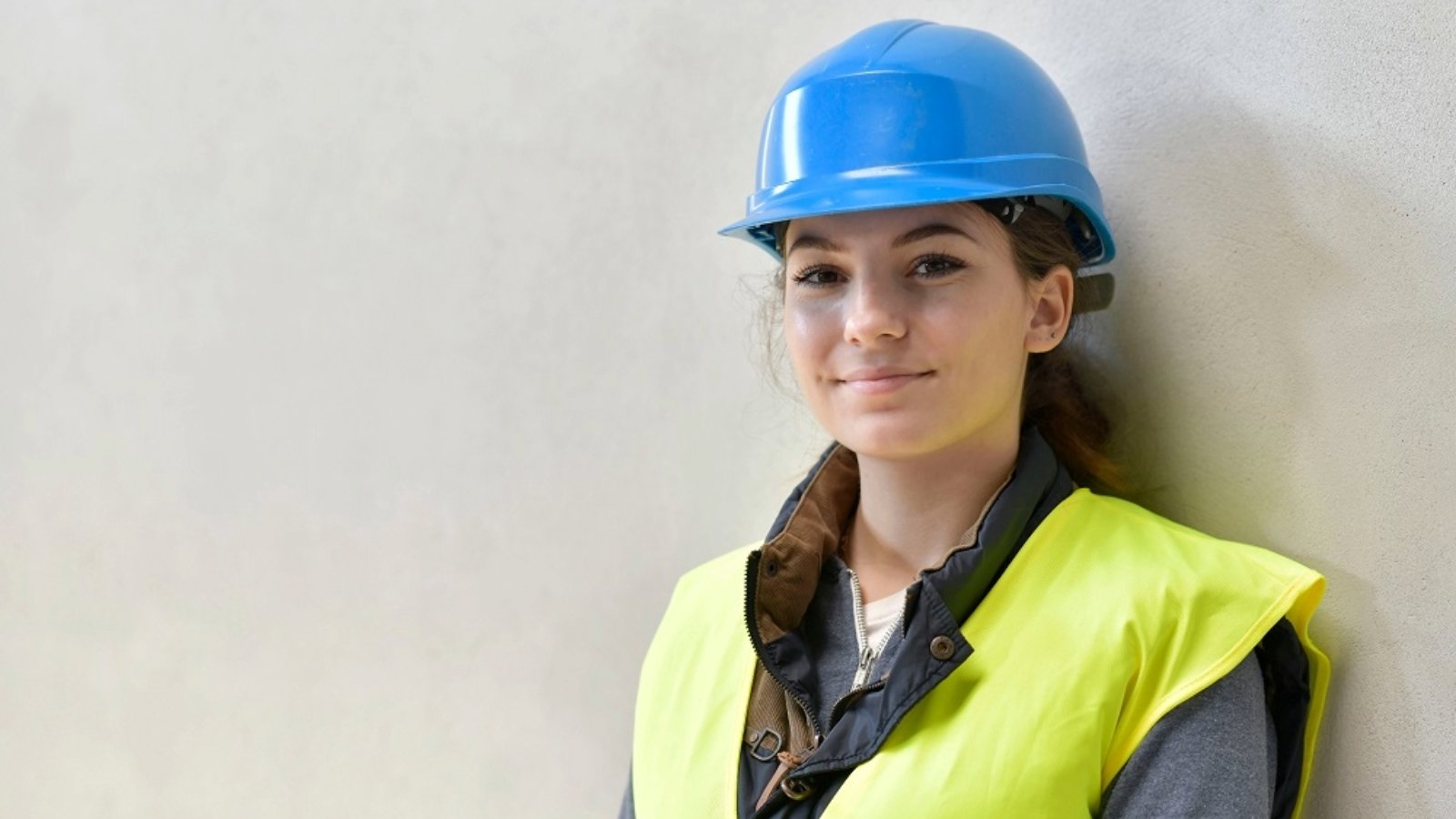 A person wearing a blue safety helmet and a high‑visibility yellow vest stands against a plain light‑coloured wall. The person is dressed in layered work clothing suitable for a construction or industrial environment.