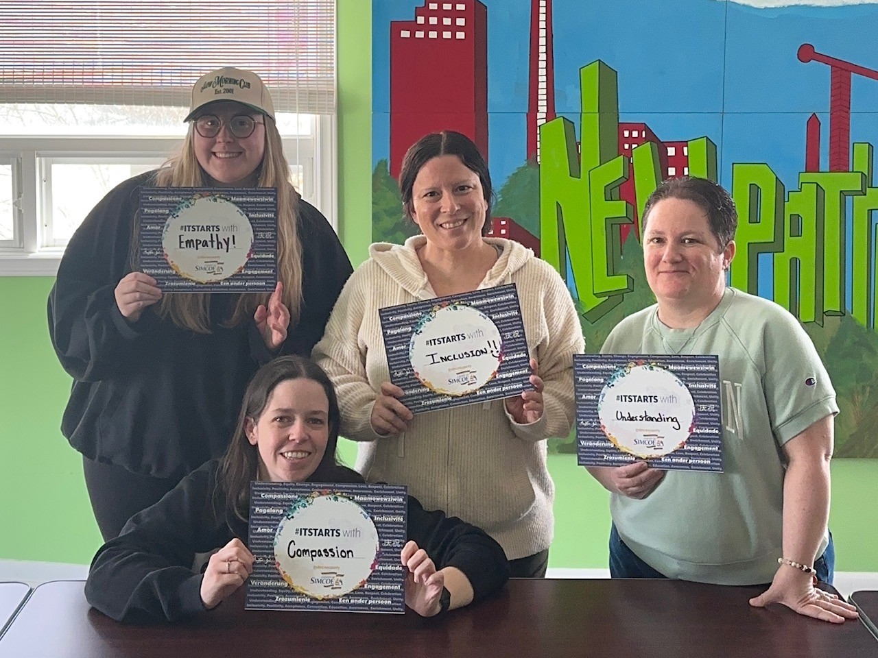 Four individuals holding certificates with words like Empathy, Inclusion, Understanding, and Compassion in a colorful room with a mural reading 'New Path'.