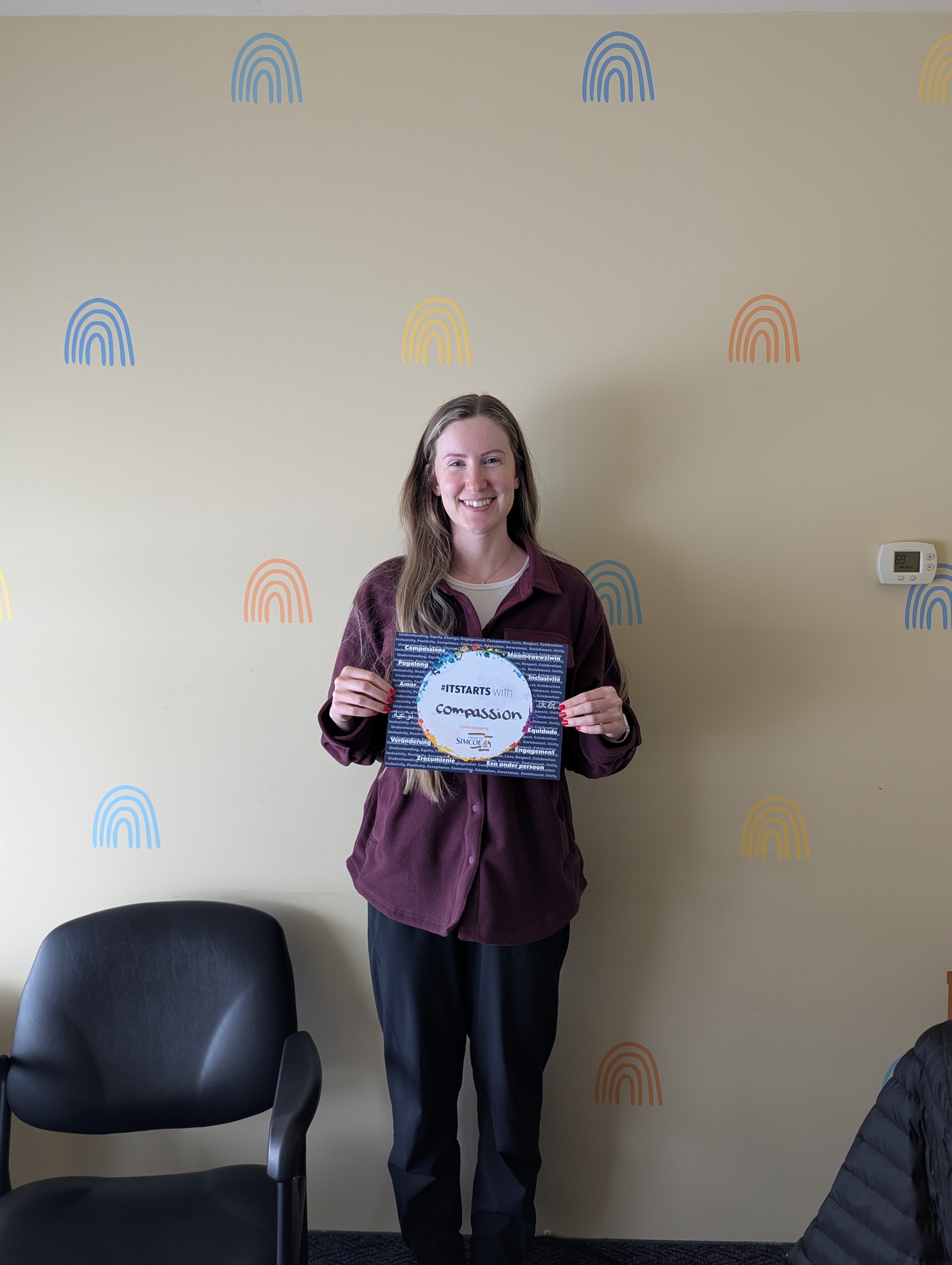 Person standing indoors holding a an ITStarts card with the word 'Compassion' against a beige wall decorated with colorful rainbow patterns.