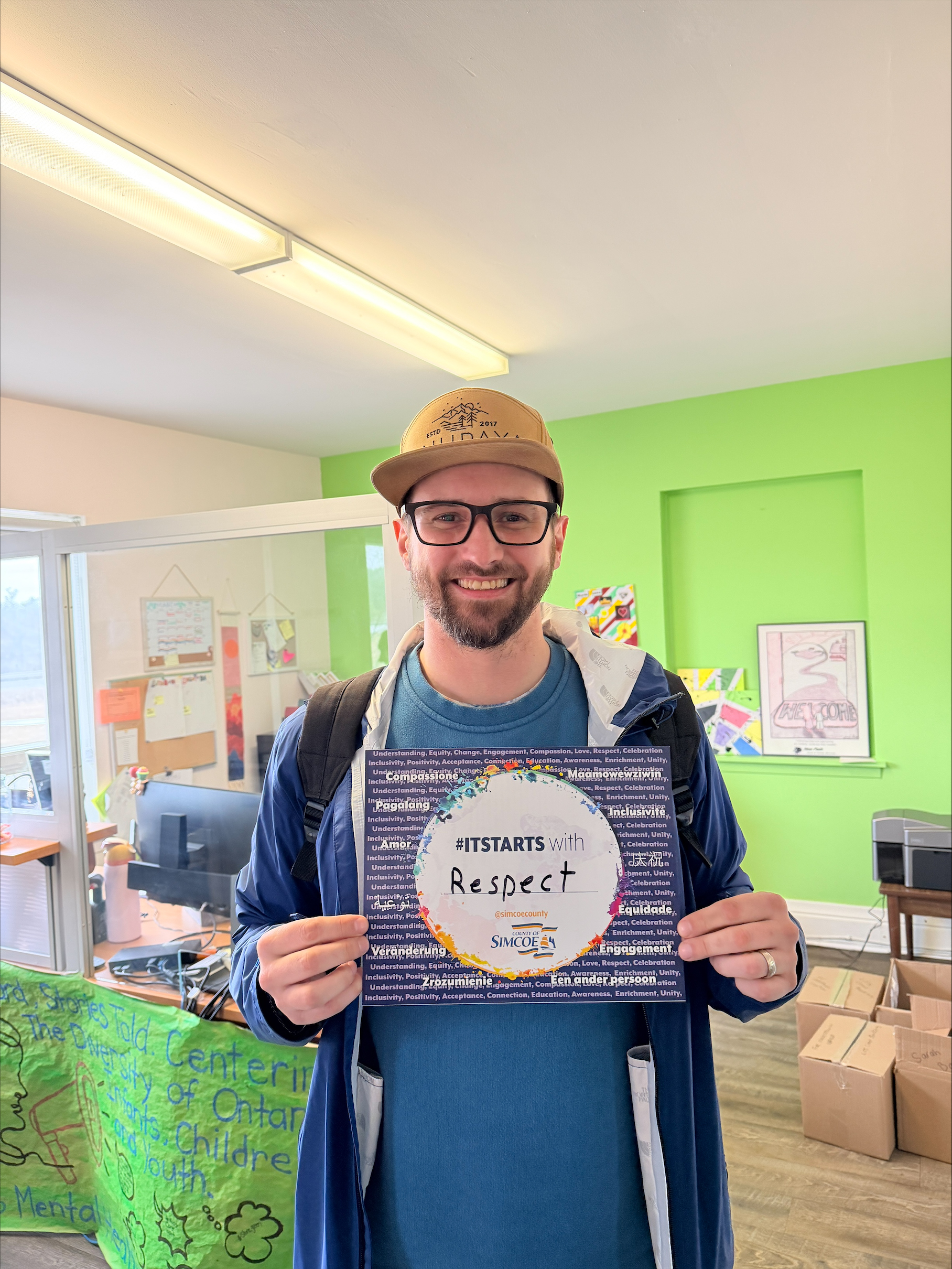 Person holding a sign that reads '#ITSTARTS with Respect' inside a brightly lit room with green walls and office supplies.