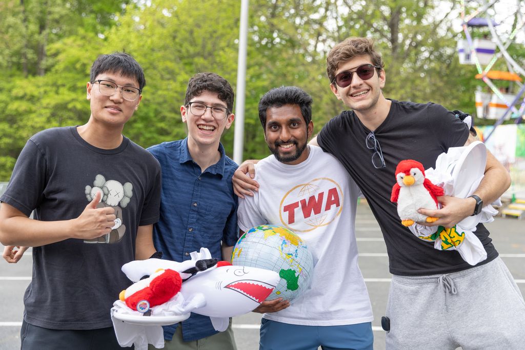 Four people stand together in a parking lot, smiling with their arms around each other. Behind them are trees and a Ferris wheel. The person on the left gives a thumbs‑up and wears a black T‑shirt with a cartoon graphic. Next to them, someone in a blue shirt and glasses smiles at the camera. The third person wears a white T‑shirt with a red “TWA” logo and holds a small globe and a plush shark. The person on the right wears a black T‑shirt and gray sweatpants, holding a red‑and‑white plush penguin and folded white clothing.