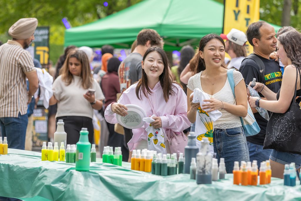 A crowd gathers outdoors around a table covered in a green cloth, where bottles of brightly colored dye are arranged for a tie‑dye activity. Two people in the foreground smile while holding plain white T‑shirts, and one also holds a frisbee printed with “Mason Day.” More attendees stand behind them chatting and holding shirts. A green canopy tent and a tall yellow banner rise in the background, creating a festive campus‑event atmosphere.