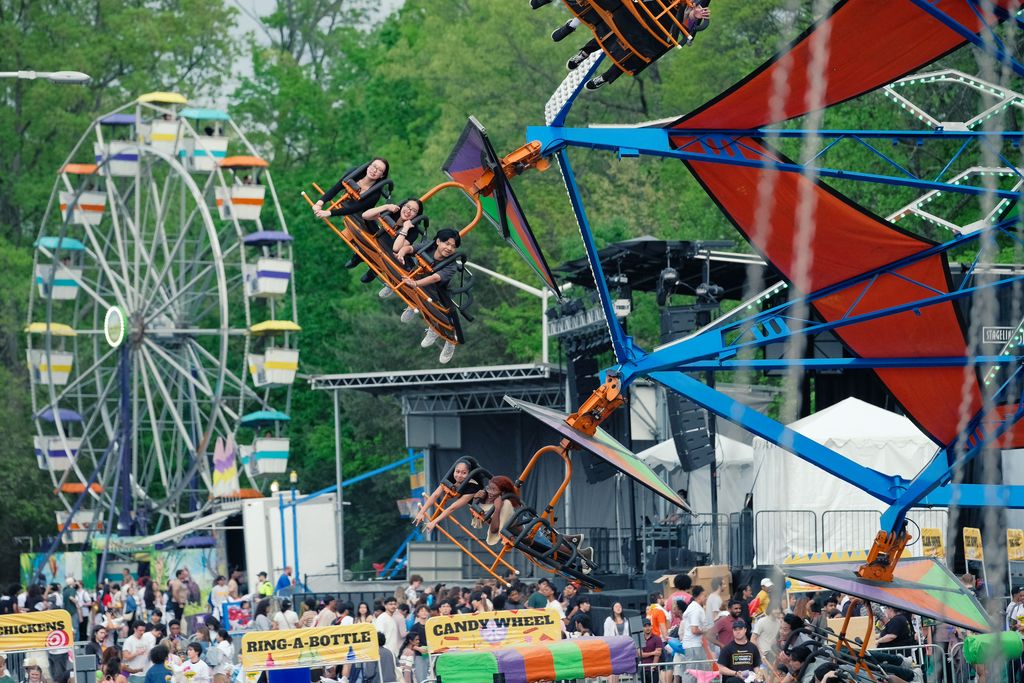 A lively carnival scene features a spinning ride with orange and black seats suspended from a blue and red structure, carrying riders through the air. Behind it, a large Ferris wheel with multicolored gondolas rises above the crowd. Below the rides, people walk among brightly decorated game booths labeled “Ring‑A‑Bottle,” “Candy Wheel,” and “Chickens.” Tents, a small stage setup, and green trees frame the festive outdoor atmosphere.