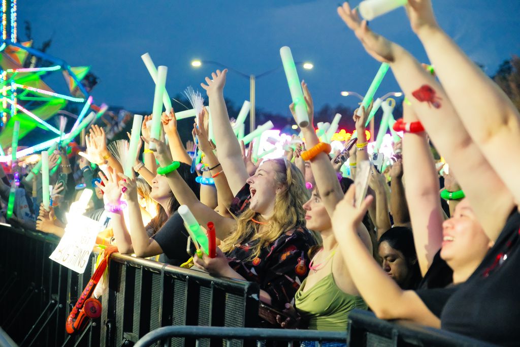 A dense crowd gathers at a nighttime outdoor event, pressed against a barricade and cheering with raised hands. Many people wave glowing foam sticks, bracelets, and other light‑up accessories that illuminate their faces in bright colors. Multicolored lights from nearby attractions or a stage cast a festive glow across the scene. One attendee holds a sign reading “WE’RE SCREAMING,” capturing the high‑energy, celebratory atmosphere.