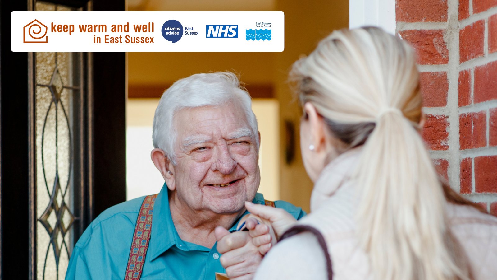 An elderly individual smiling at a visitor at their doorway under a sign that says 'Keep warm and well in East Sussex' with NHS and local council logos.