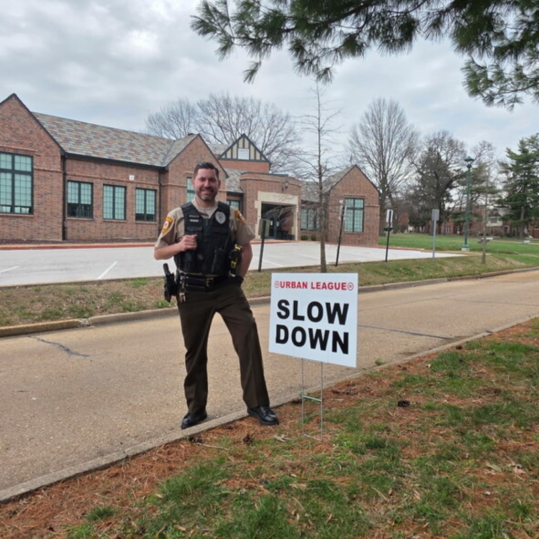 Uniformed officer standing next to a roadside sign that reads 'Urban League SLOW DOWN' in a suburban neighborhood.