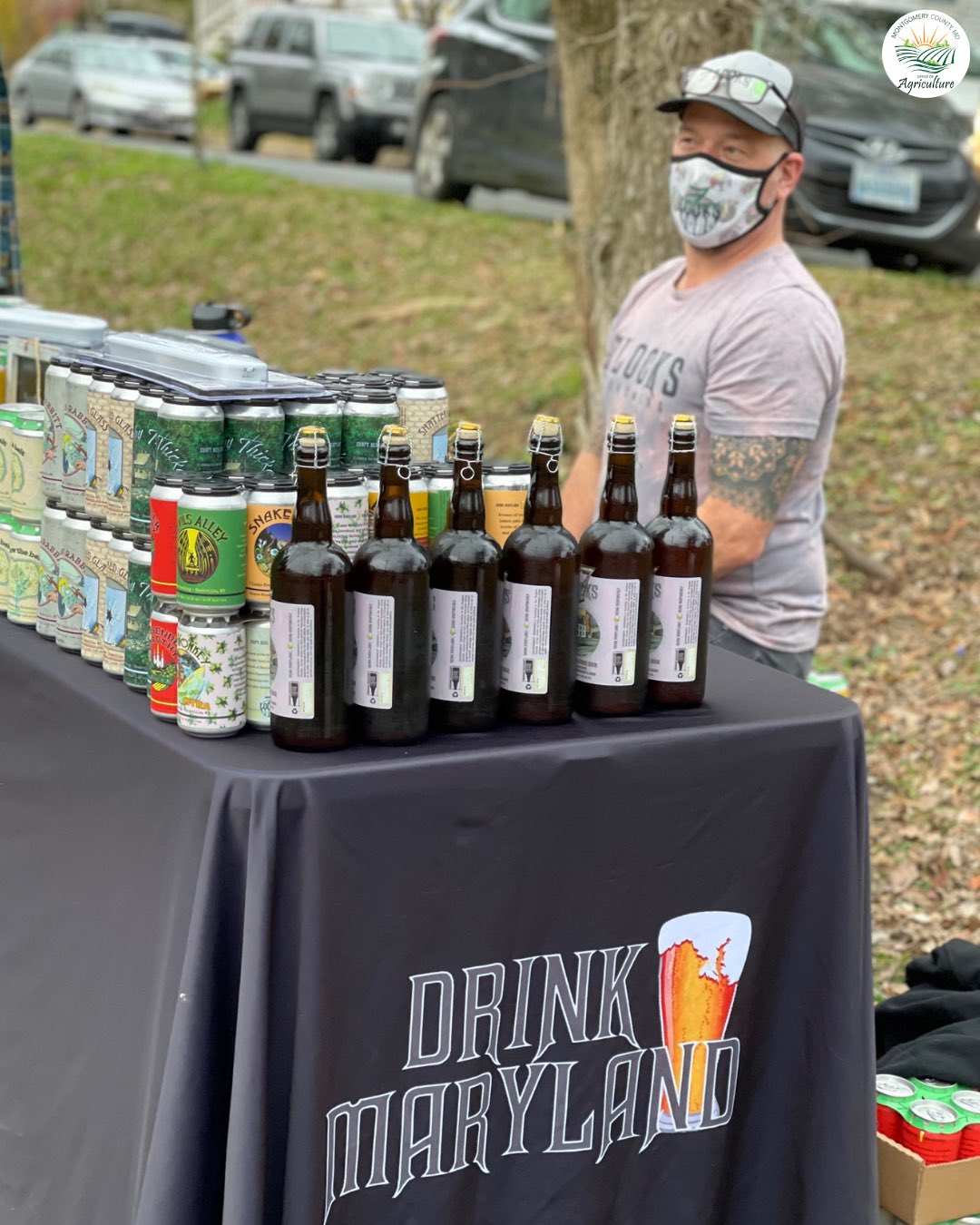 A vendor standing behind a table displaying bottles and cans of craft beverages at an outdoor farmers market. Montgomery County Office of Agriculture logo included.