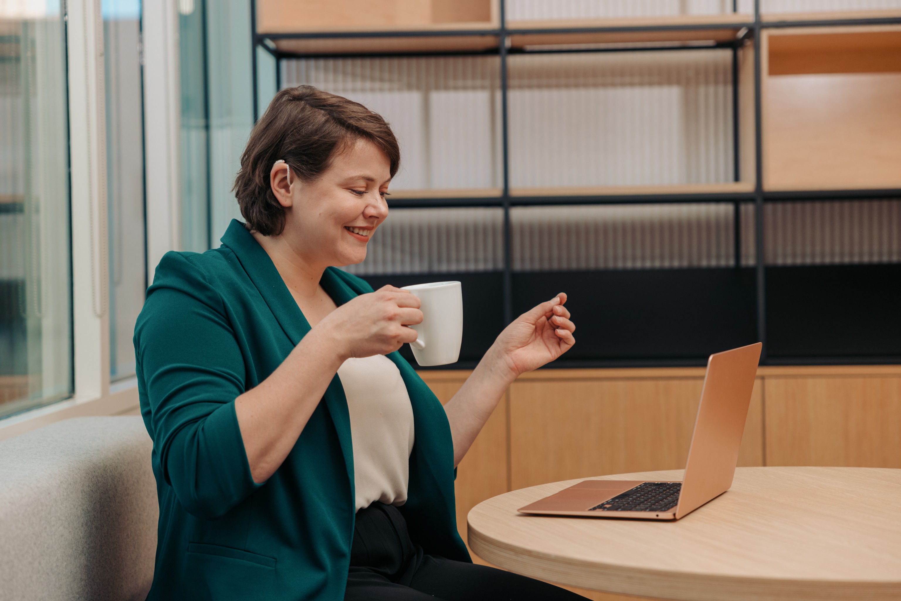 Professional in a Virtual Meeting
A professional works on their laptop while holding a cup of coffee in an office. They have their hair tucked behind their ear to show off their hearing aid.

Photo Credit: Disability:IN