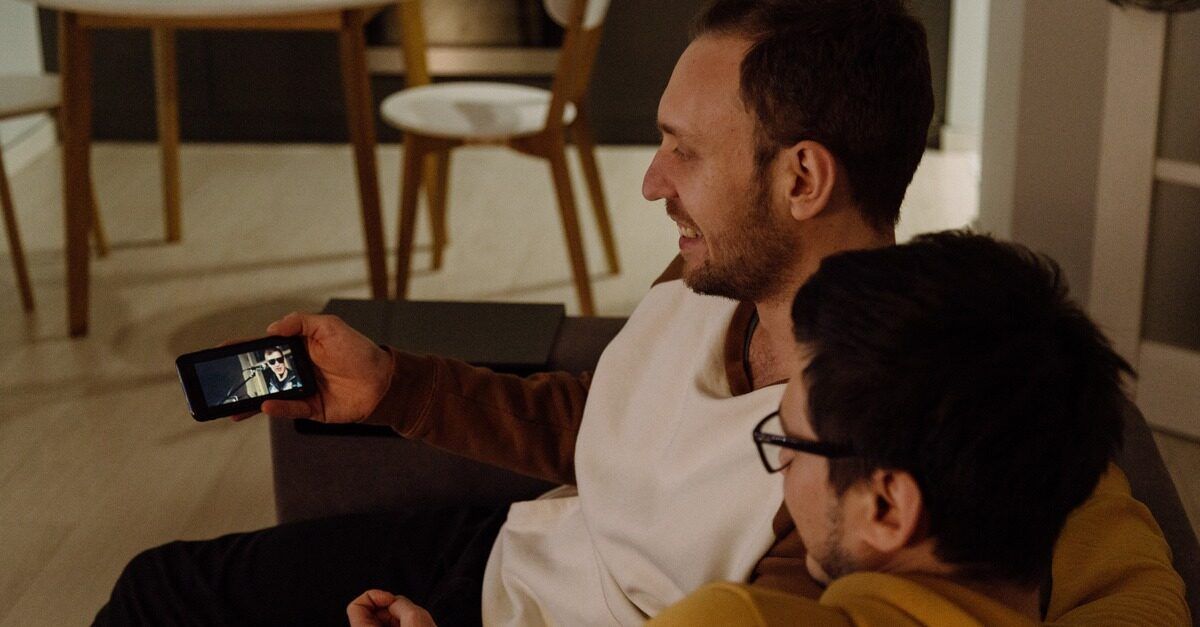 Two dark-haired men watch a video on a smartphone while sitting on a couch.