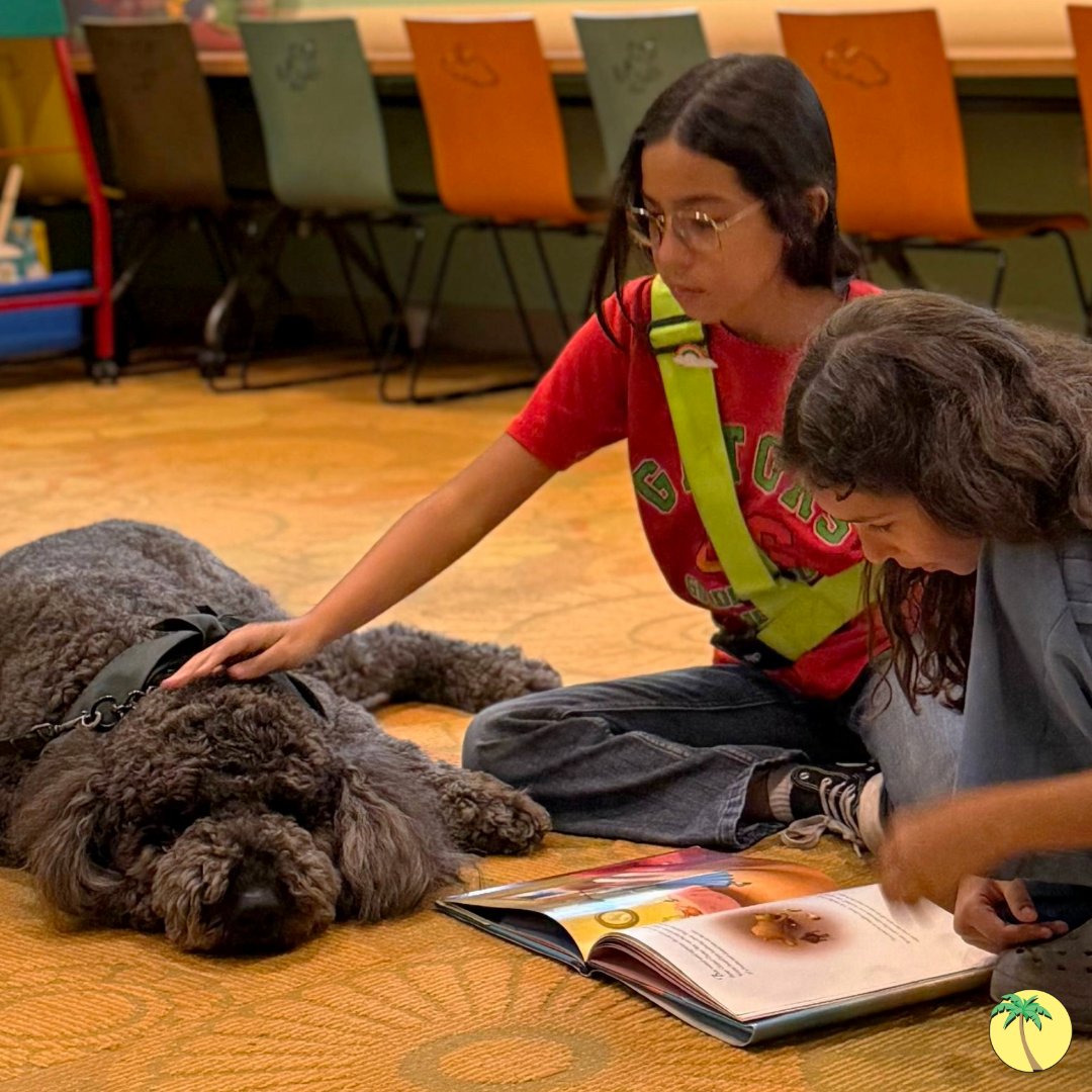 A girl with a safety patrol vest and her friend sitting on the floor and reading a book, while Reggie the police dog lays comfortably beside them. The girl with the safety patrol vest is petting Reggie while they read.