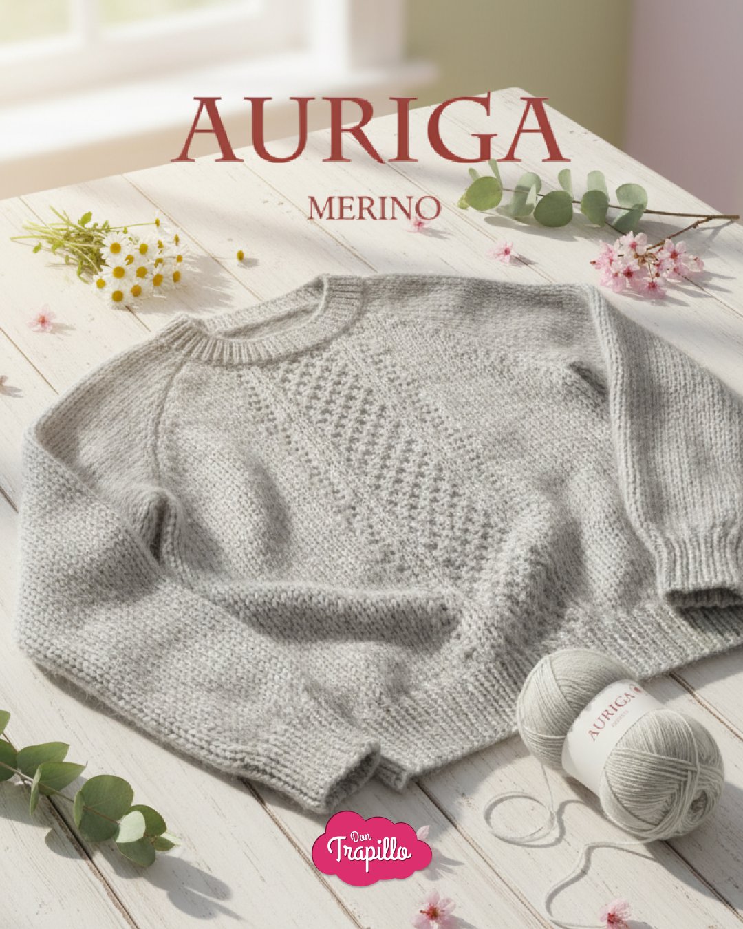 Jersey gris de lana merino con ovillos etiquetados "Auriga", sobre mesa blanca decorada con flores.