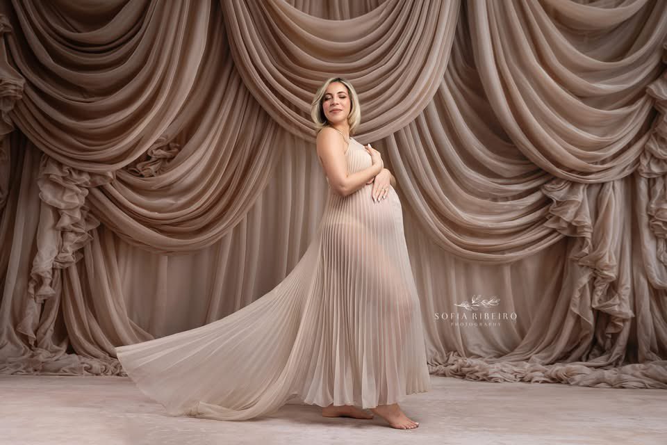 Elegant maternity photoshoot featuring a woman in a flowing neutral gown posed in front of a champagne-colored draped fabric backdrop with soft cascading folds.