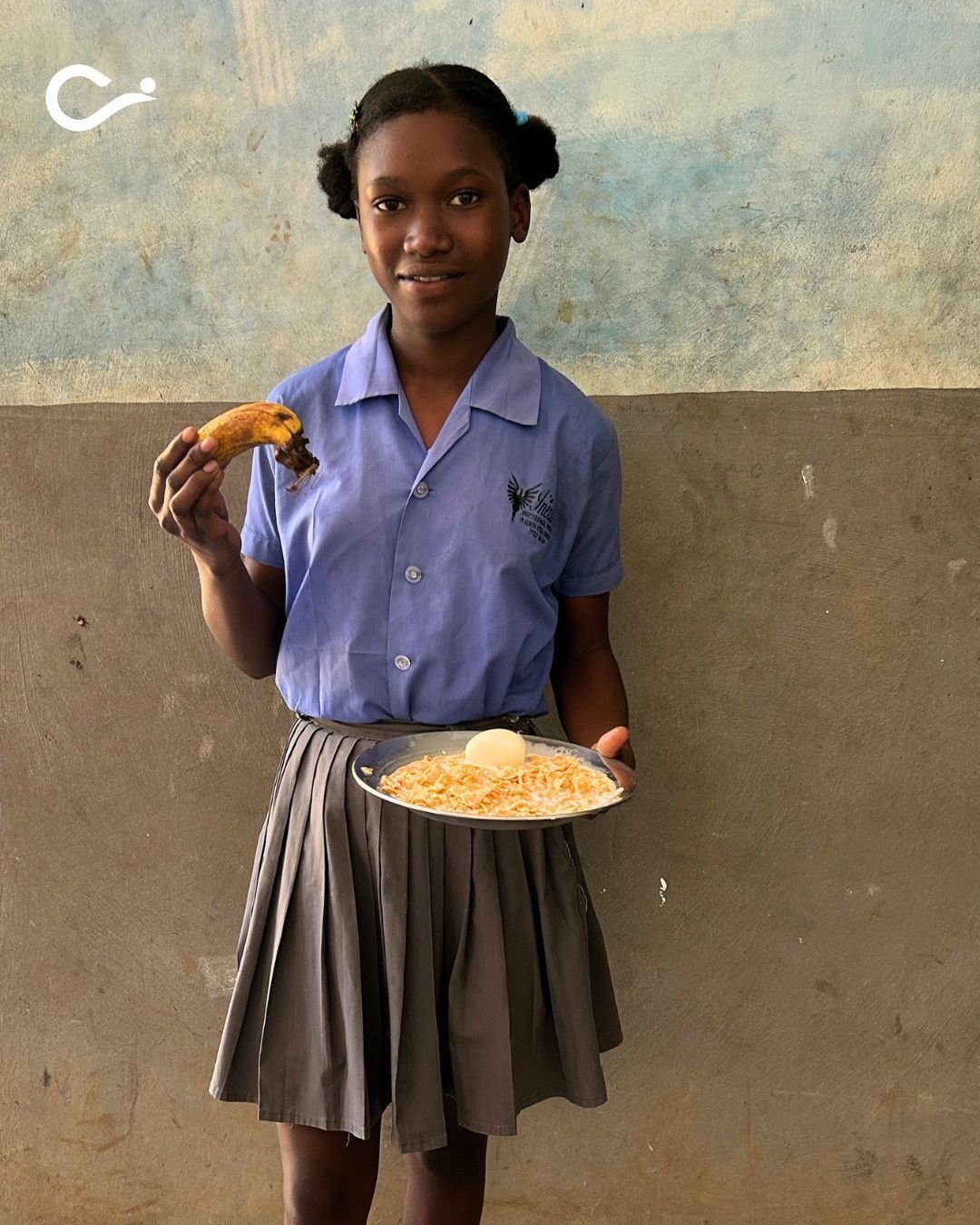 A girl stands against a wall holding a plate of food in one hand and a piece of fruit in the other.