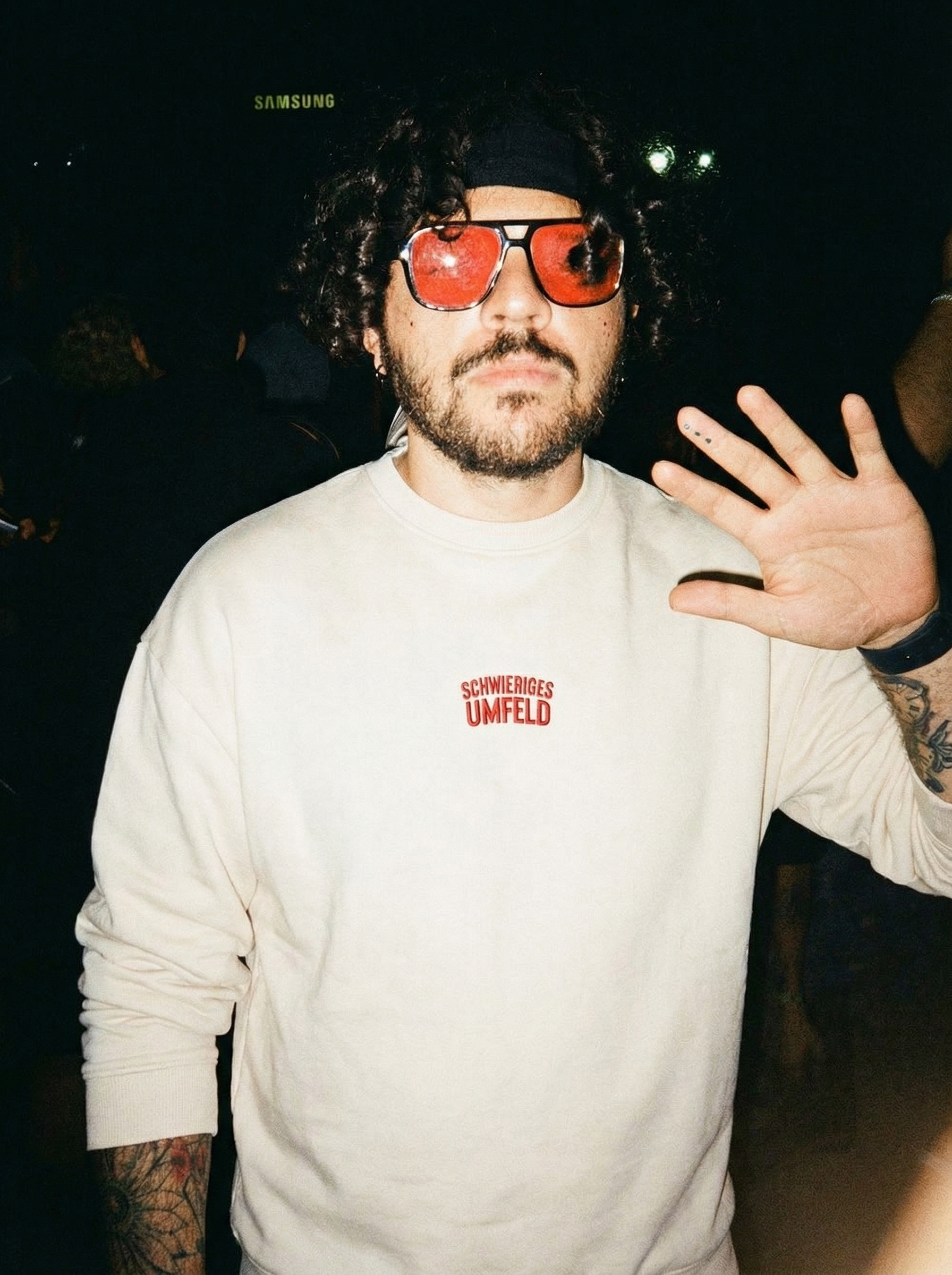 Mann mit 'schwieriges Umfeld' Sweatshirt im Club hält seine Hand Richtung Kamera.