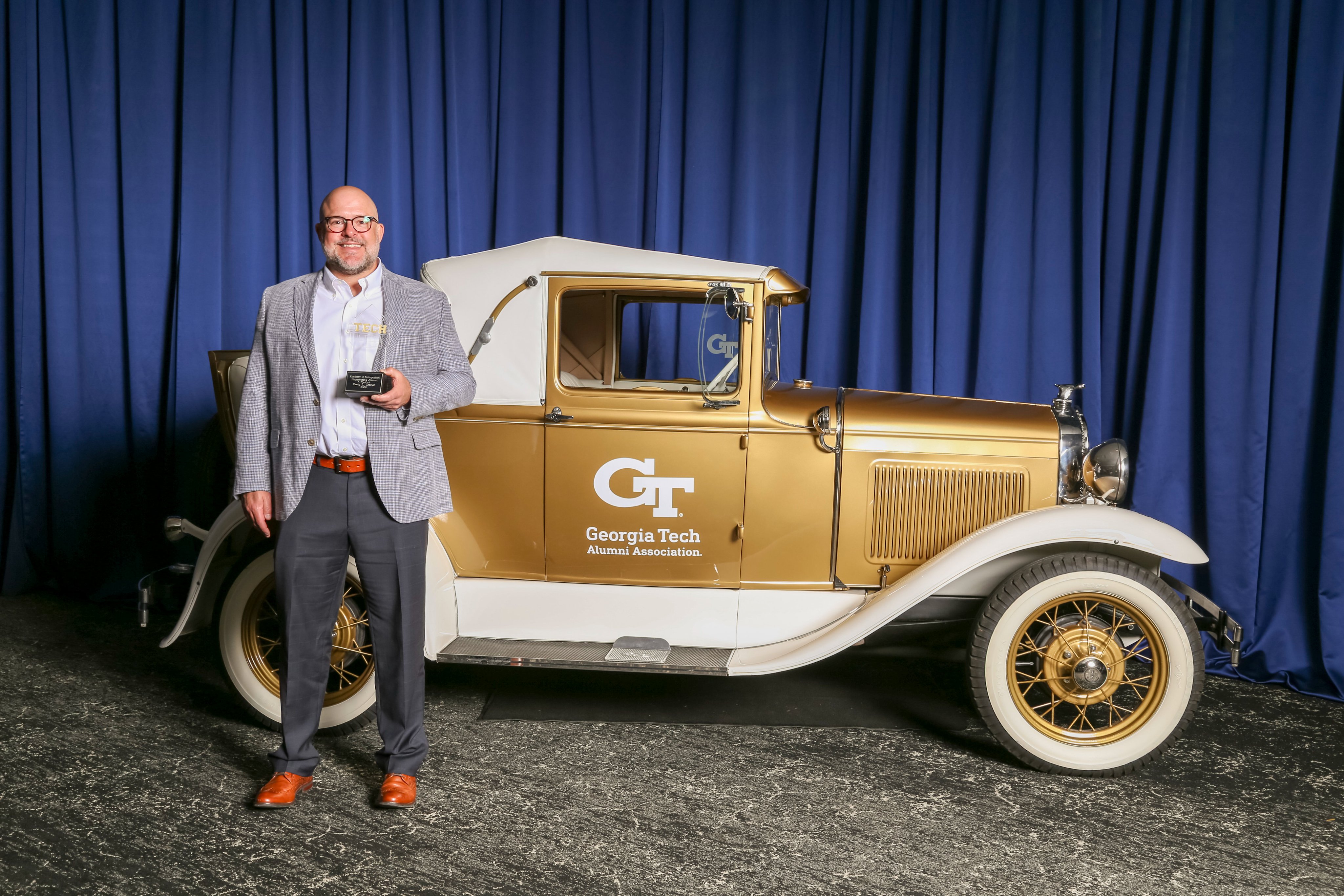A person stands beside the Georgia Tech Alumni Association Ramblin’ Wreck car, holding a glass award, with blue curtains in the background.