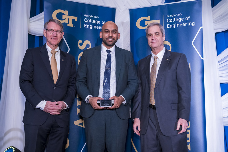 Three people stand on a stage in front of Georgia Tech College of Engineering banners; the person in the center holds a glass award.