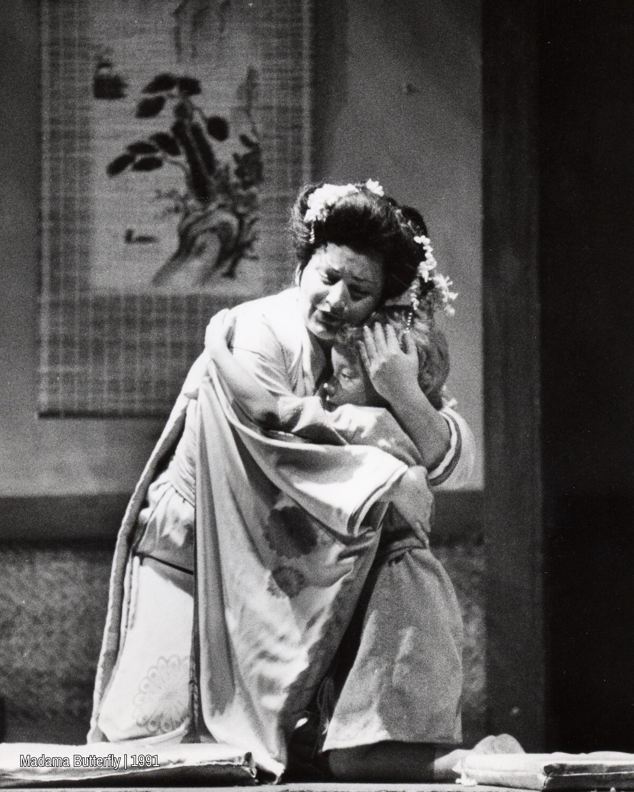 Black and white image of a performer kneeling and embracing a child, both held close in an intimate, emotional moment on stage.