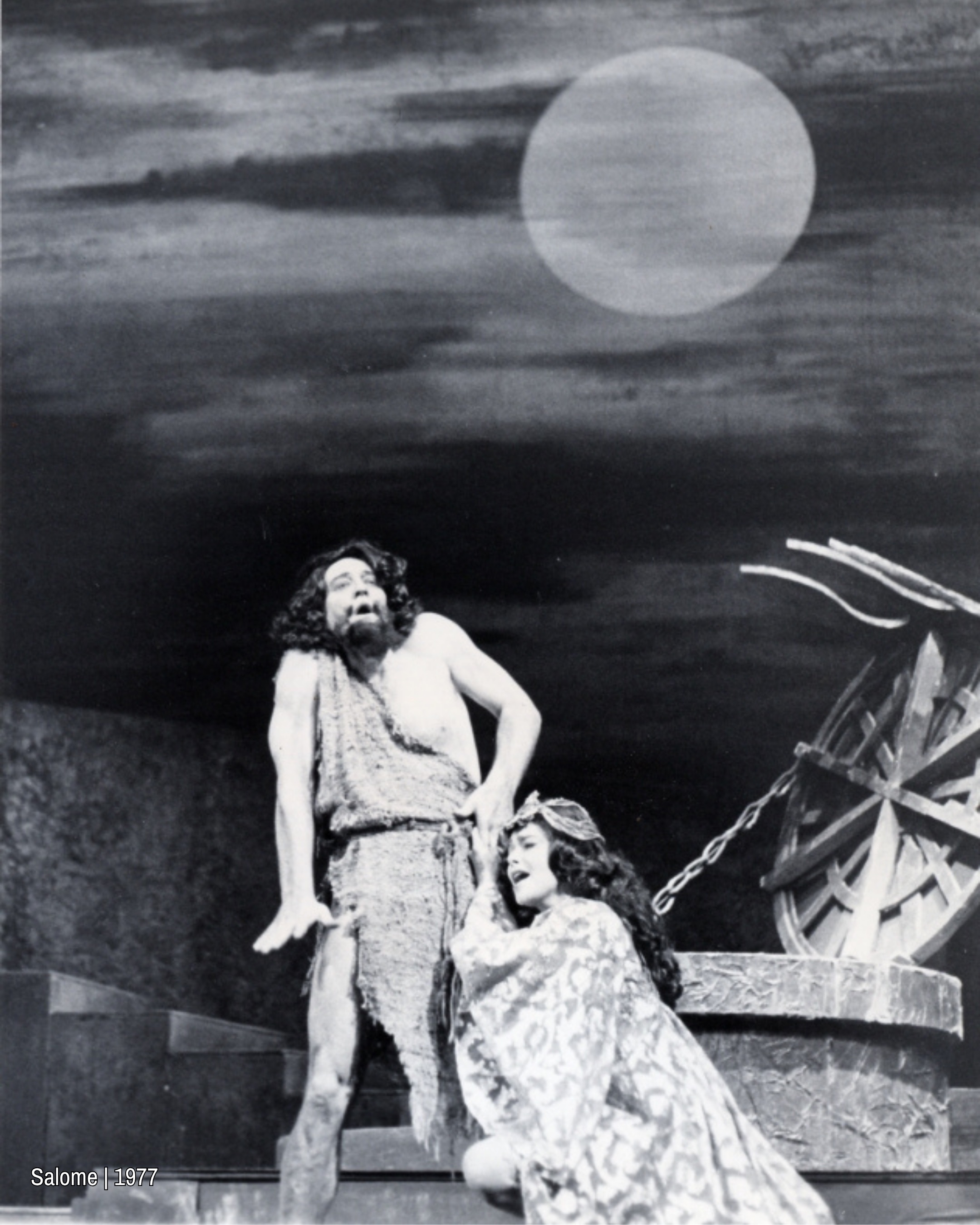 Black and white image of two performers on a stark stage, one standing and the other kneeling beside them, both expressing dramatic emotion under a large moon backdrop.
