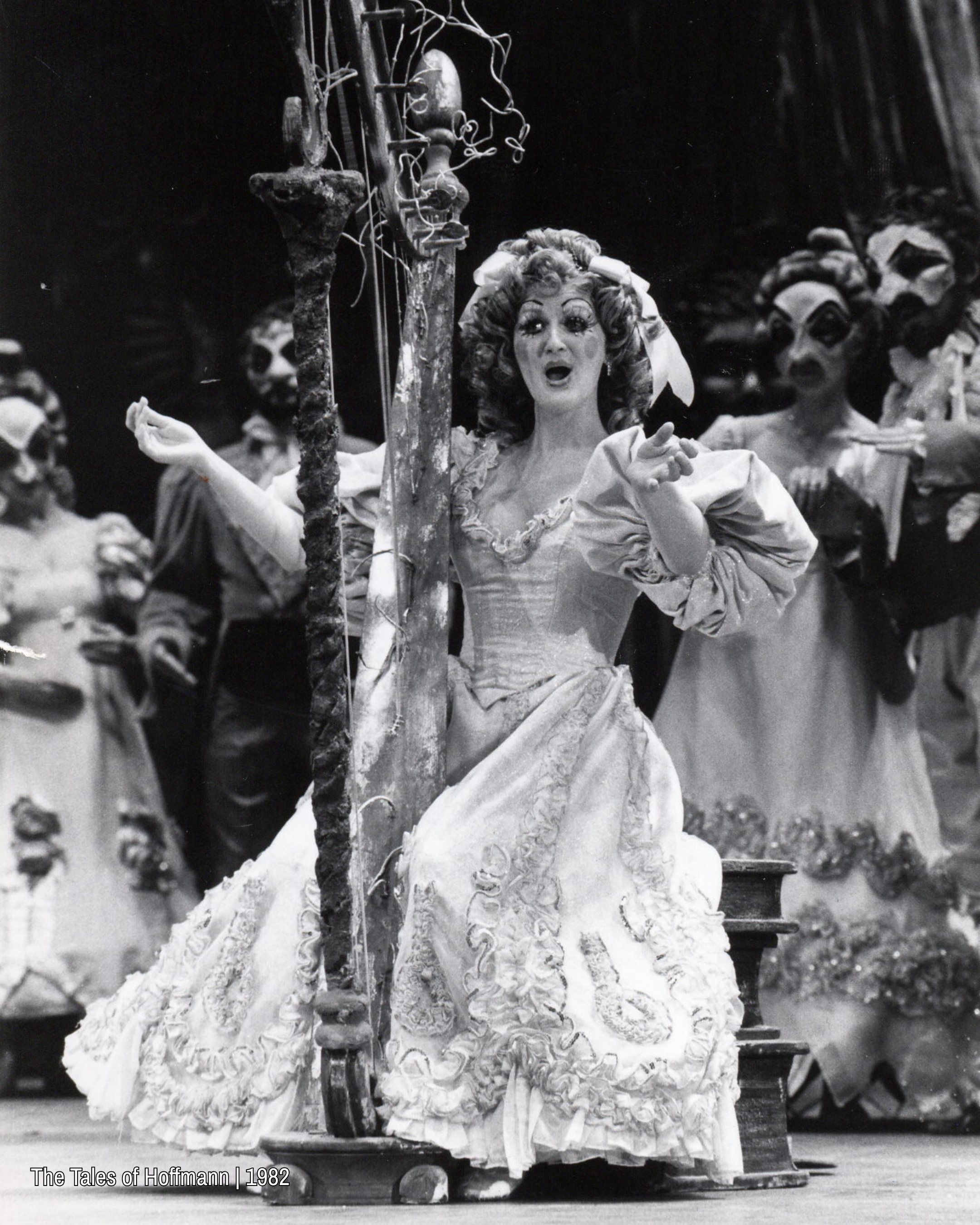 Black and white archival photo of an opera singer in an elaborate 19th century costume standing centre stage, singing with arms open. Other performers in theatrical costumes and masks stand behind her in a dimly lit set.