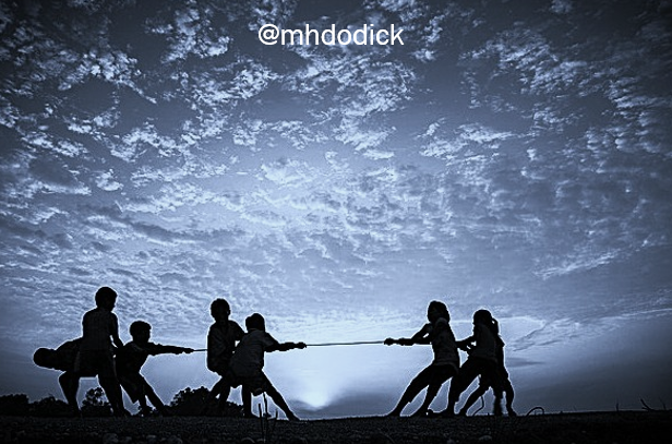 Image in a blue hue of two groups of children seen in silhouette participating in a tug of war. The sun is setting against a cloud-filled sky. At the top centre: @mhdodick