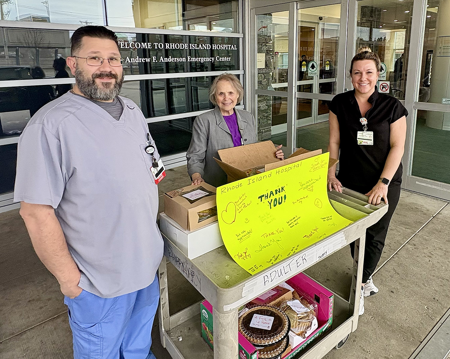 Three people stand outside the entrance to Rhode Island Hospital’s emergency center beside a cart filled with donated baked goods, including pies and cakes. A bright yellow poster reading “Thank you!” with handwritten messages is propped on the cart. 