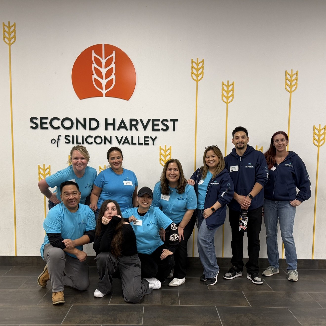 A group of Mirastar volunteers wearing blue shirts and jackets pose in front of a wall with the "Second Harvest of Silicon Valley" logo. Some are standing and others kneeling, smiling at the camera.