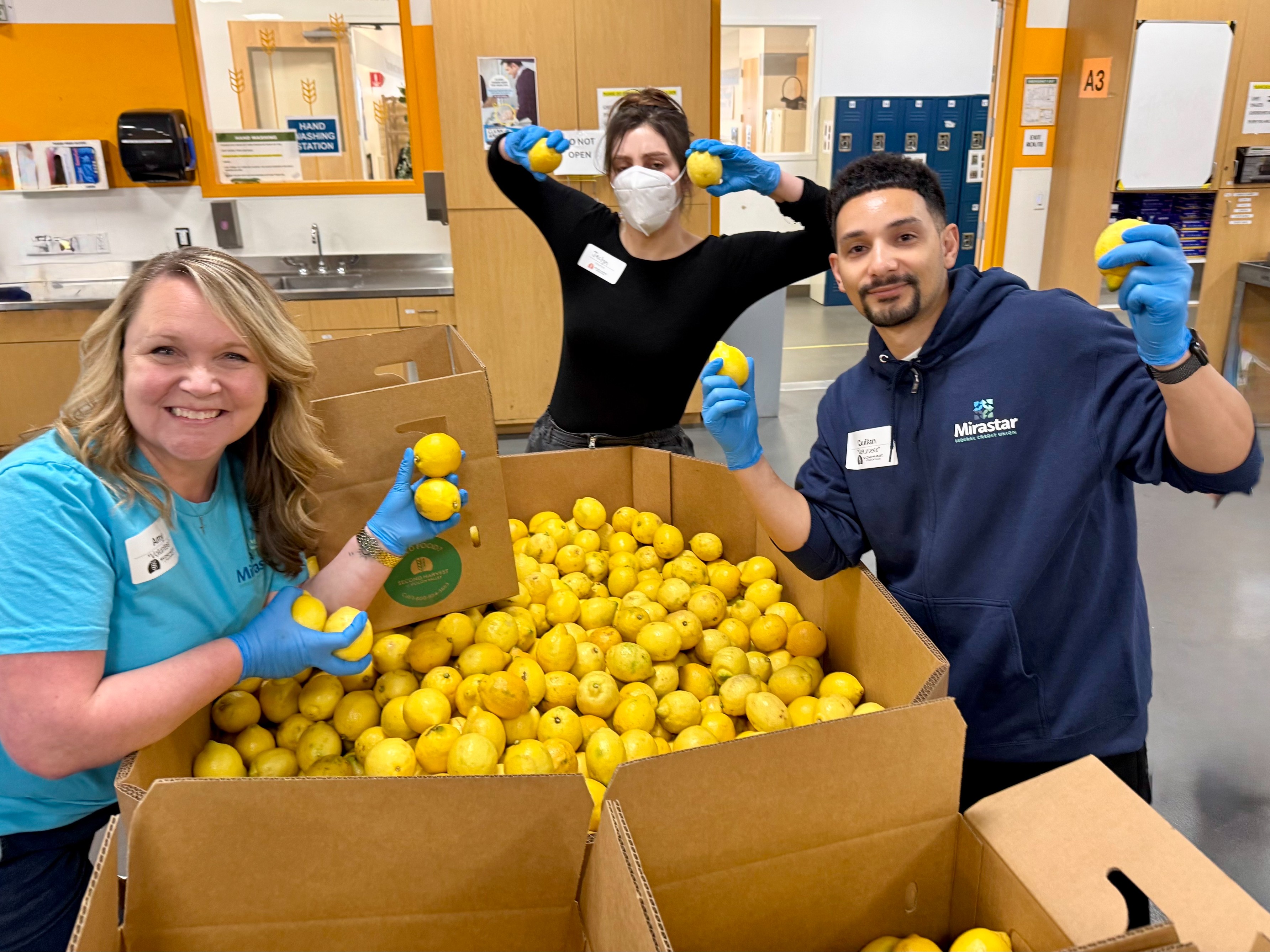 Three Mirastar volunteers in a room are smiling and holding lemons over a large box filled with lemons. They are wearing gloves and casual clothing. The person on the left is wearing a blue T-shirt with "Mirastar" written on it. Cabinets and lockers are visible in the background.