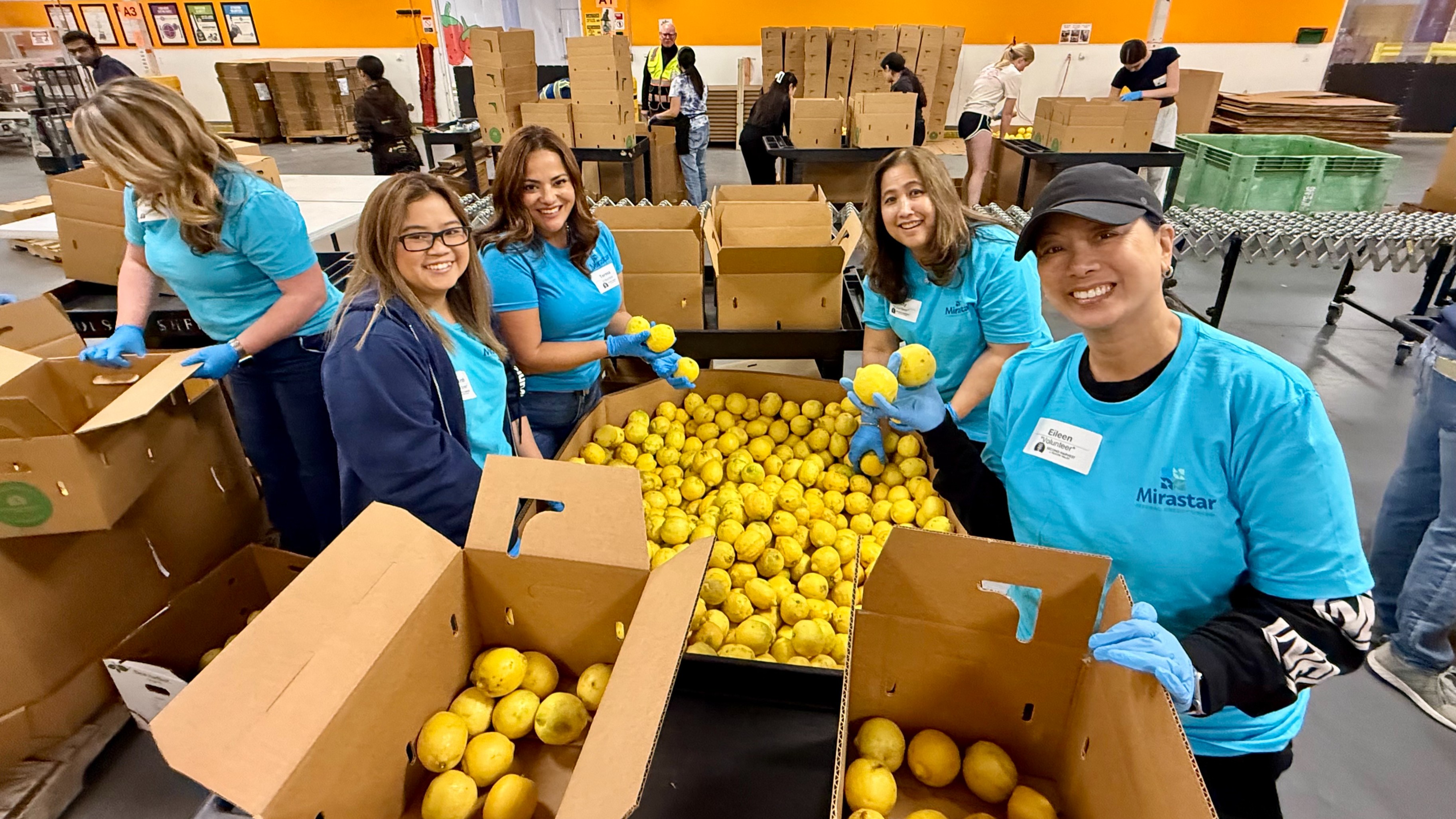 A group of Mirastar volunteer in blue Mirastar shirts are sorting lemons into boxes in a warehouse setting. Visible are stacks of cardboard boxes and green crates. The background includes more workers and shelved items.