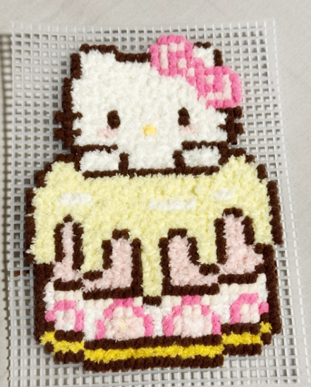 Alt Text: A handmade textured pixel art piece of a Hello Kitty cake on a white plastic mesh grid. The artwork is crafted using a tufting or punch needle technique, giving it a soft, fuzzy appearance. Hello Kitty, wearing her signature pink bow, is perched atop a multi-layered cake featuring dripping pale-yellow frosting. The bottom layers show pink and dark brown patterns, all rendered in a thick, plush, and pixelated style.