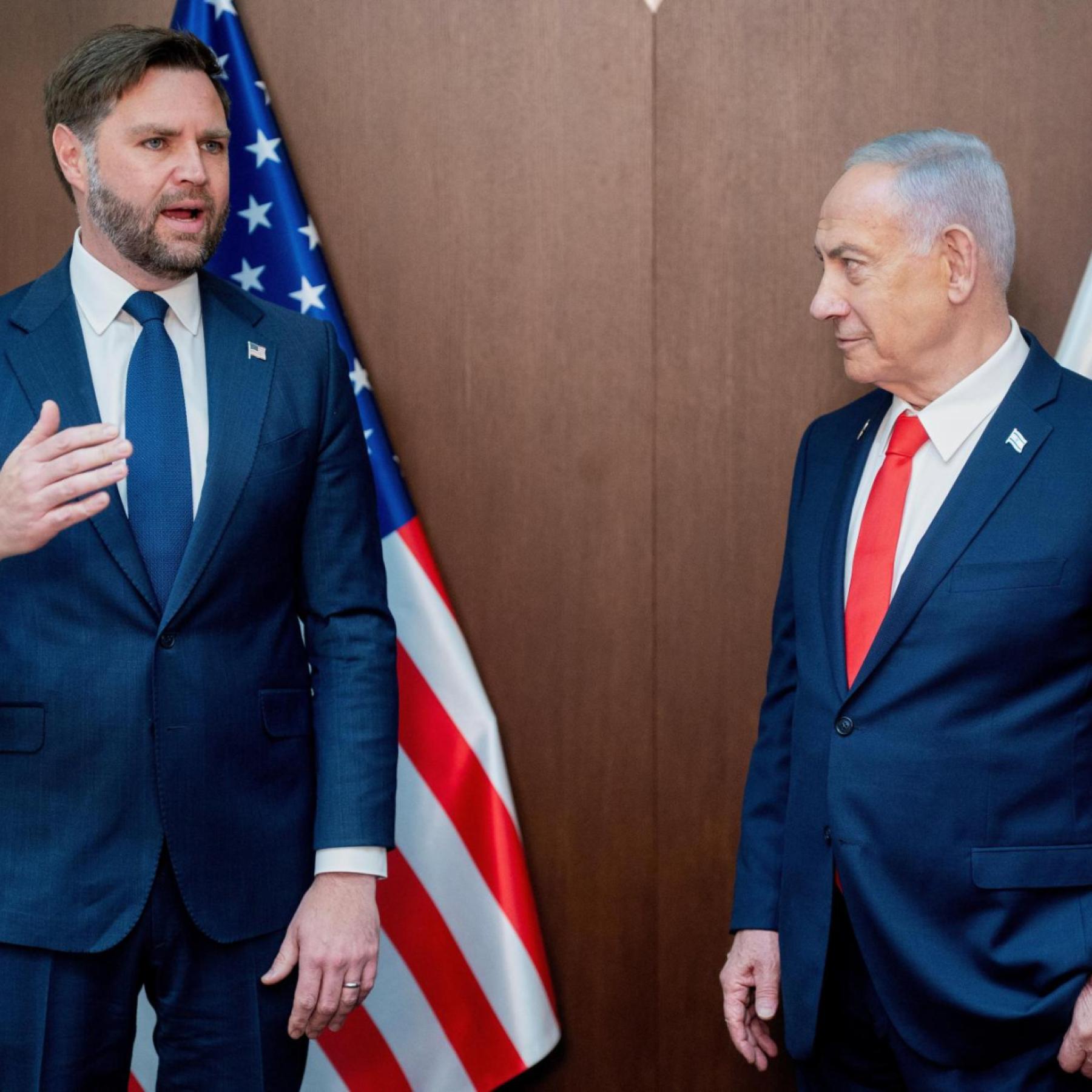 D Vance, thick beard and intense stare, stands in a dark suit shaking hands with Benjamin Netanyahu—white hair neatly combed, navy jacket, red tie—both facing a wall of microphones and flashing cameras. American and Israeli flags hang behind them, smiles forced and tight, like a diplomatic deal sealed under pressure. Pure tension in every frame.