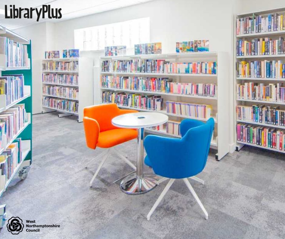 Bright library corner with orange and blue chairs around a small round table surrounded by white bookshelves filled with books.