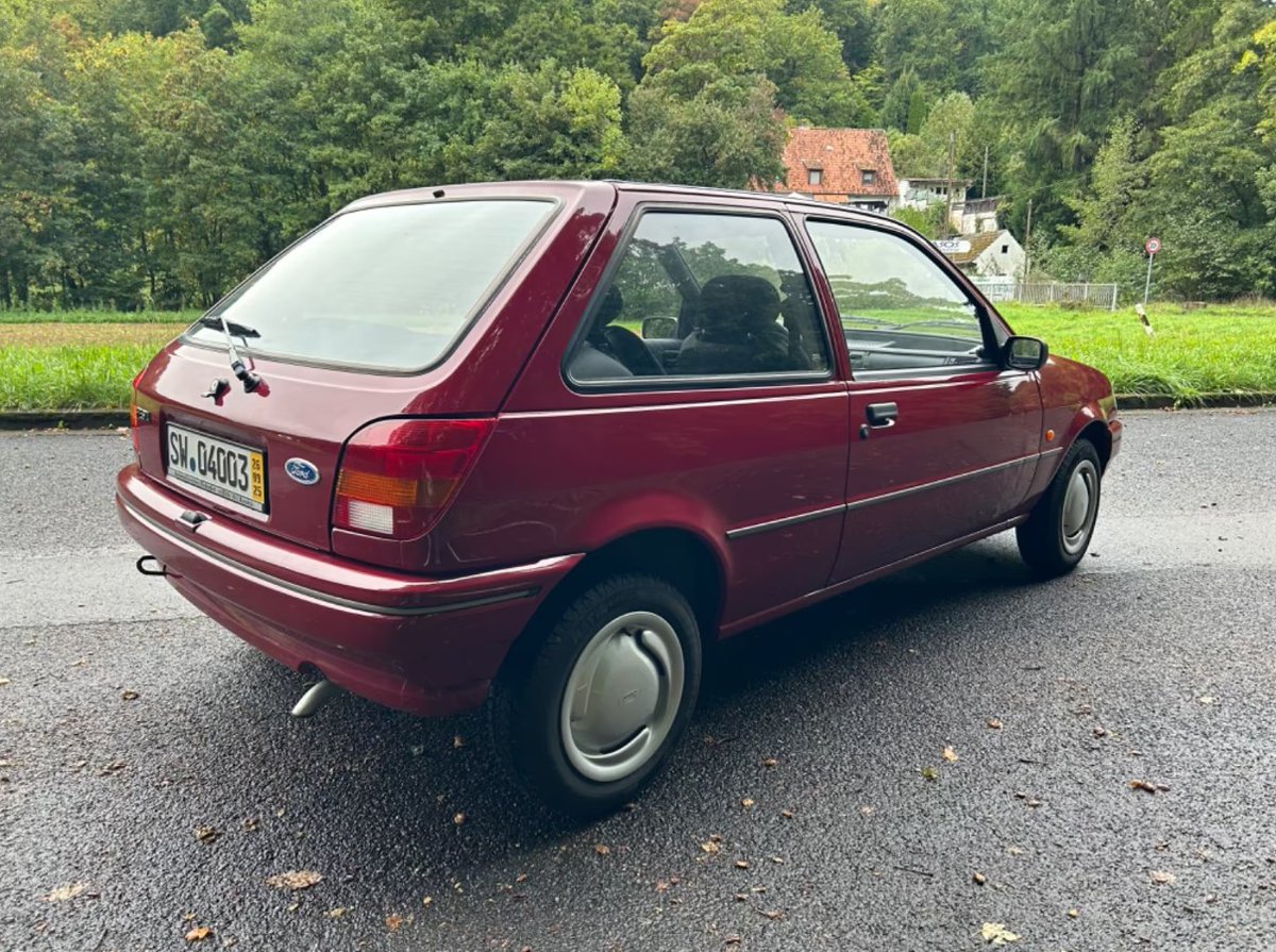 Der rote Dreitürer von halb hinten: charakteristische Rückleuchten in Spindelform, die an der oberen äußeren Ecke knapp unterhalb der Gürtellinie in eine an der Seite entlang laufenden Sicke übergehen, Heckklappe mit Scheibenwischer, glatte Radkappen mit organischer Formgebung