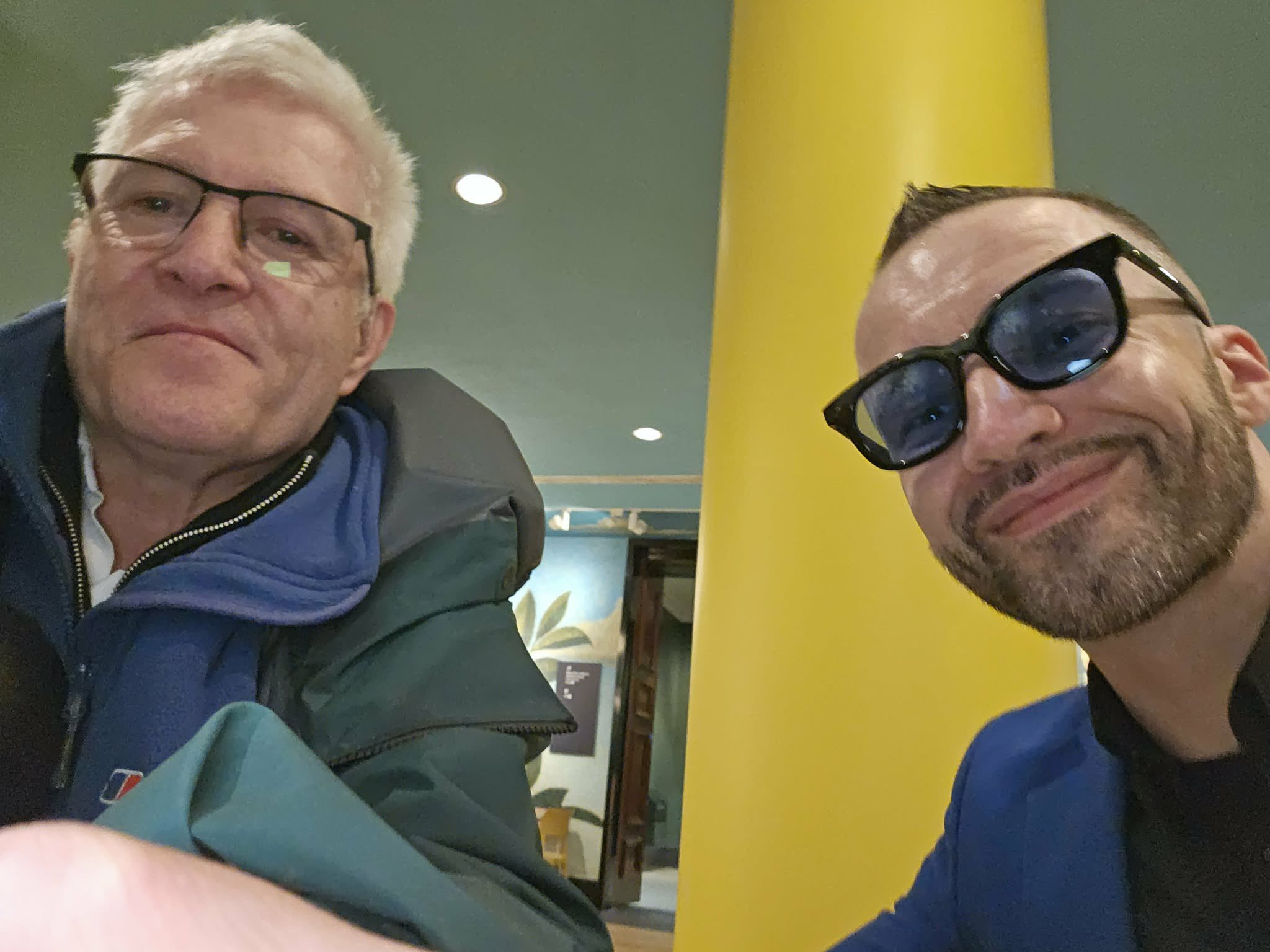 Man with white hair wearing glasses and a green and blue coat sits next to a man wearing sunglasses
