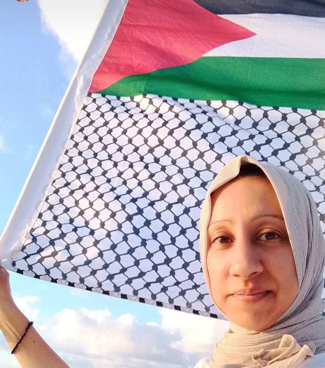 Photo of a woman in hijab (Dr. Zaheera Soomar) holding a Palestinian flag with a keffiyeh patterned border, with blue sky + clouds in the background, 