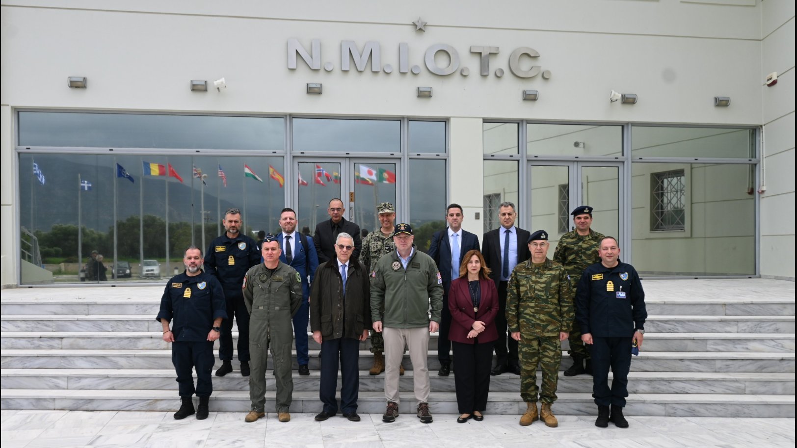 Image, slightly cropped, from: https://x.com/NMIOTC/status/2036404945541288121, showing a group of fourteen people, civilian & military, a lone woman among them, standing in front of a building

The relevant Tuesday 24 March 2026 @NMIOTC tweet reads:
"🇺🇸 On Thursday, 19 March 2026, the Permanent Representative of the United States to #NATO, H.E. Ambassador Matthew G. Whitaker visited NMIOTC in the context of his official visit to  🇬🇷.
"@USAmbNATO @Hellenic_MOD @GreeceNATO @USEmbassyAthens"

NMIOTC:
"NATO Maritime Interdiction Operational Training Centre (NMIOTC) is a training facility for NATO, at Crete Naval Base, in Souda Bay, Crete, Greece.
"Located at the Northern Sector of the base (Marathi), it enables the NATO forces to better execute surface, sub-surface, aerial surveillance, and special operations activities that are necessary for the support of Maritime Interdiction Operations (MIO)."
https://en.wikipedia.org/wiki/NATO_Maritime_Interdiction_Operational_Training_Centre
+++