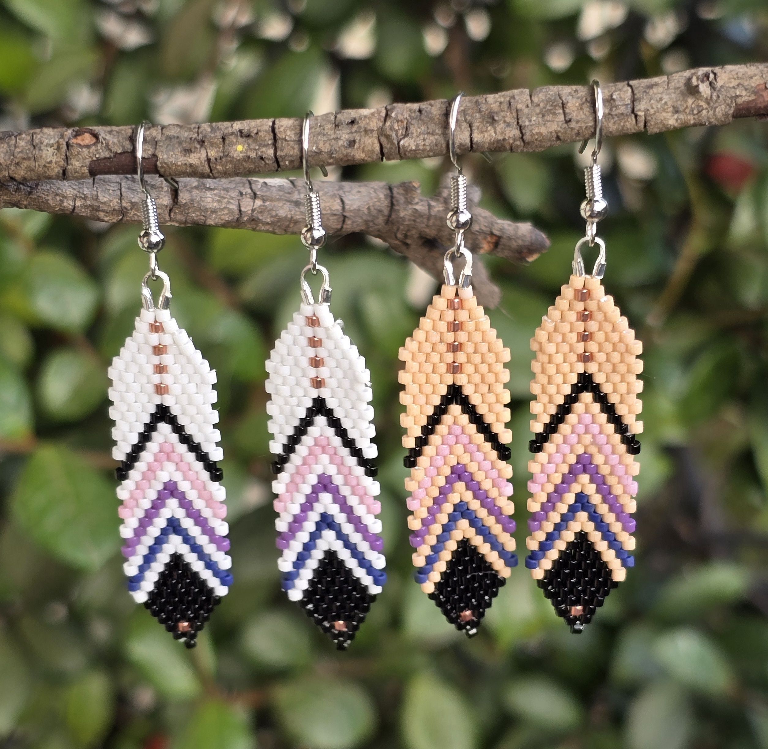 beaded feather earrings, one pair white background, the other light tan, shades of black, blue, purple, pink, rose gold, background of natural greenery