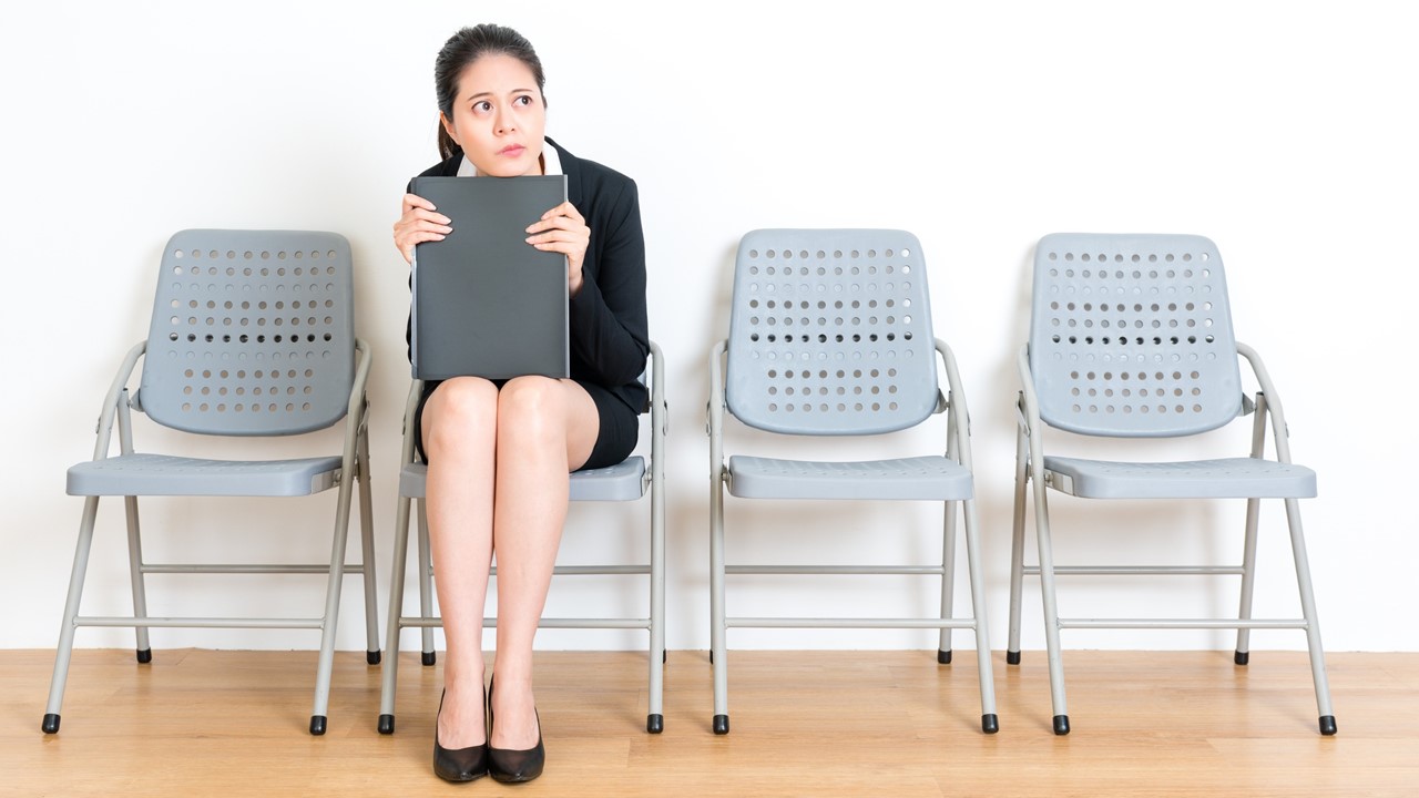 Woman wearing a suit and sitting on a row of seats, holding a folder and looking nervous