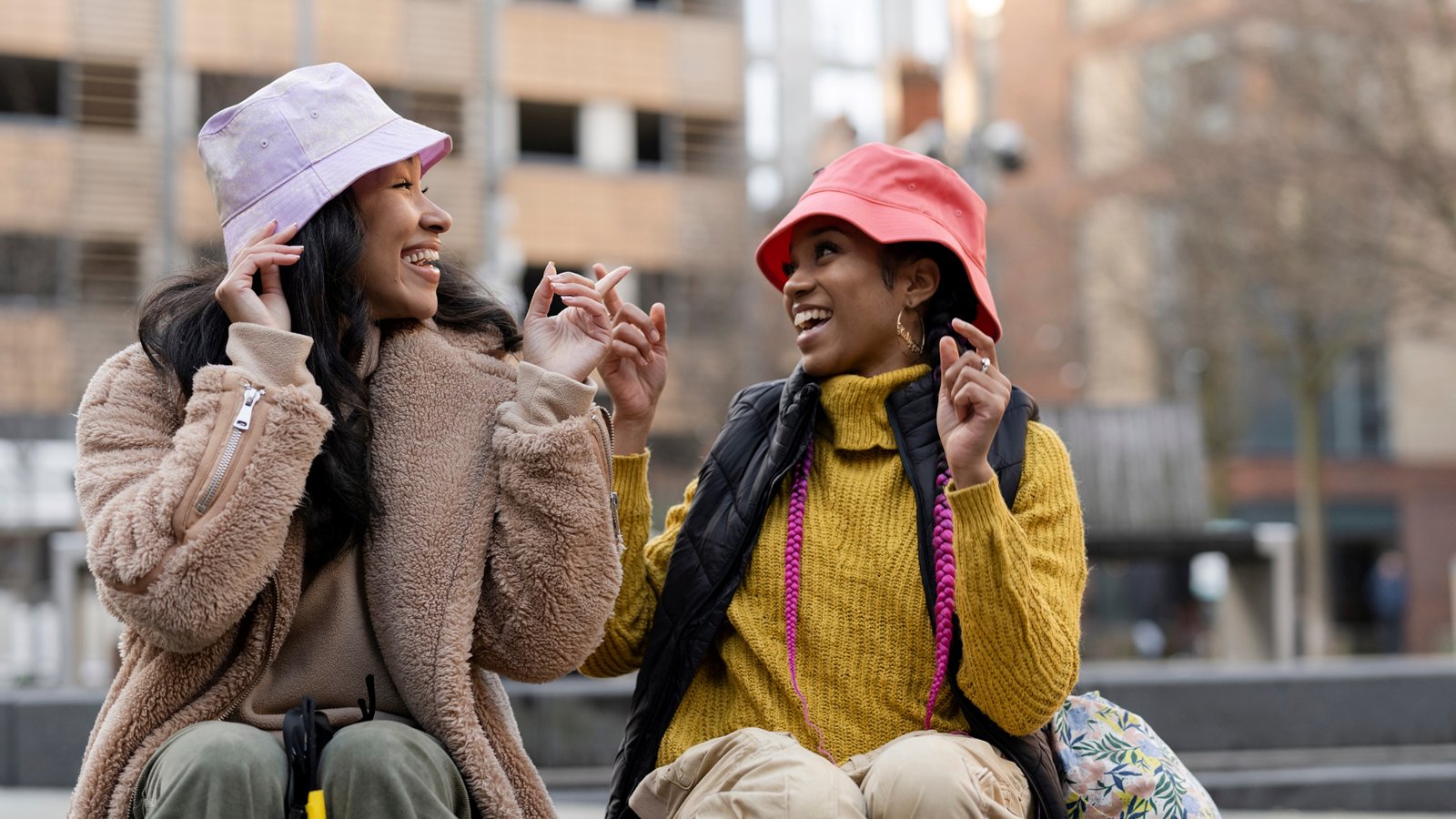 Two people sitting outdoors in an urban setting, facing each other and holding hands in a playful gesture. Both are wearing casual clothing with colourful knitwear and bucket hats—one in a light purple hat and beige fleece jacket, the other in a bright red hat and mustard yellow sweater with a dark vest. Behind them are modern buildings and a few leafless trees, suggesting a cool season.