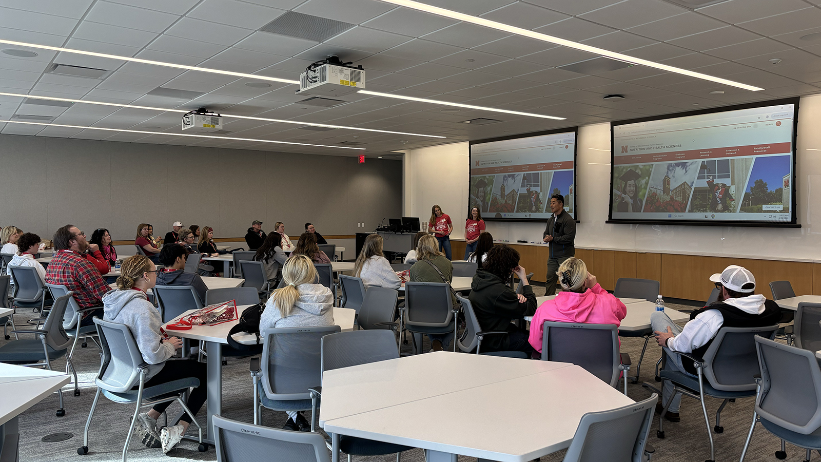 a faculty member and two student ambassadors stand at the front of a classroom speaking to visitors sitting at tables