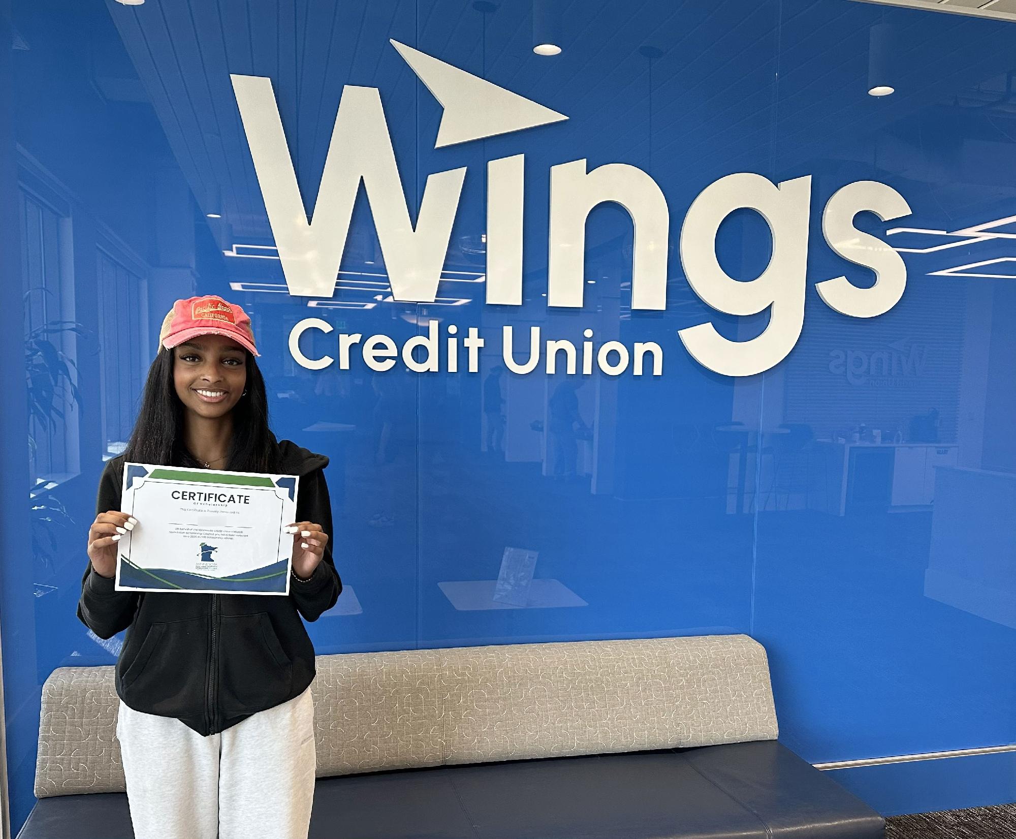 A person is standing in front of a blue wall with the Wings Credit Union logo. They are holding a certificate and wearing a red cap. There's a couch in the foreground.